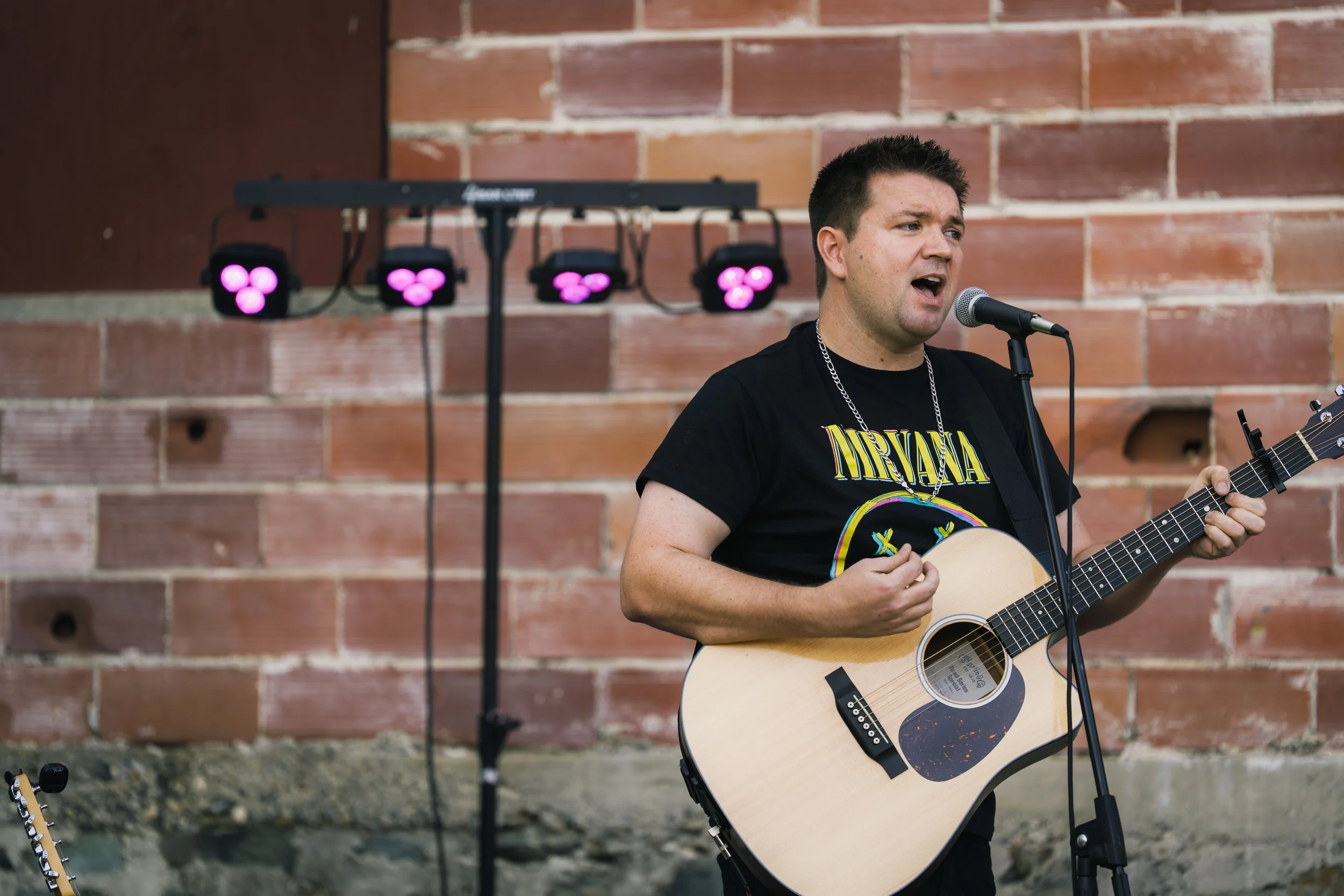 A man wearing a Nirvana t-shirt is singing into a microphone and playing an acoustic guitar, standing against a brick wall with purple stage lights.