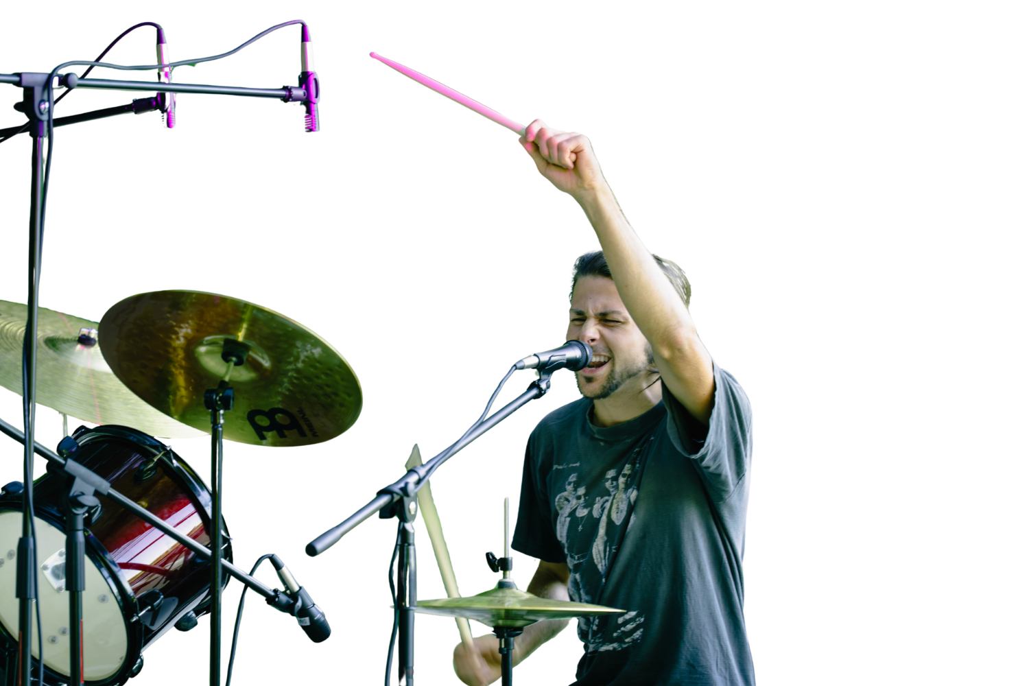 A man with dark hair and a beard singing into a microphone, holding a drumstick in one hand, with drums and cymbals around him, performing passionately.