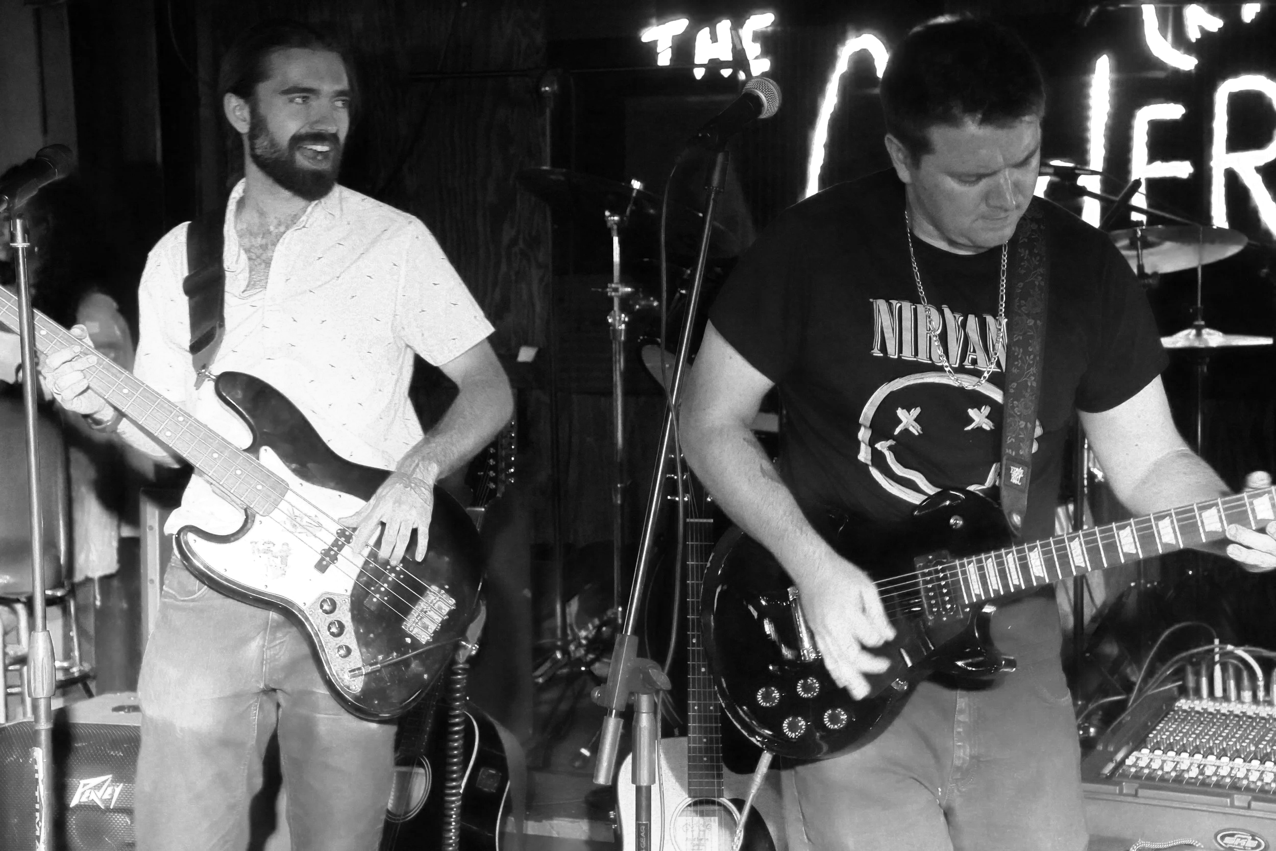 Two male musicians performing on stage with guitars in a bar or club setting, with a neon sign in the background.