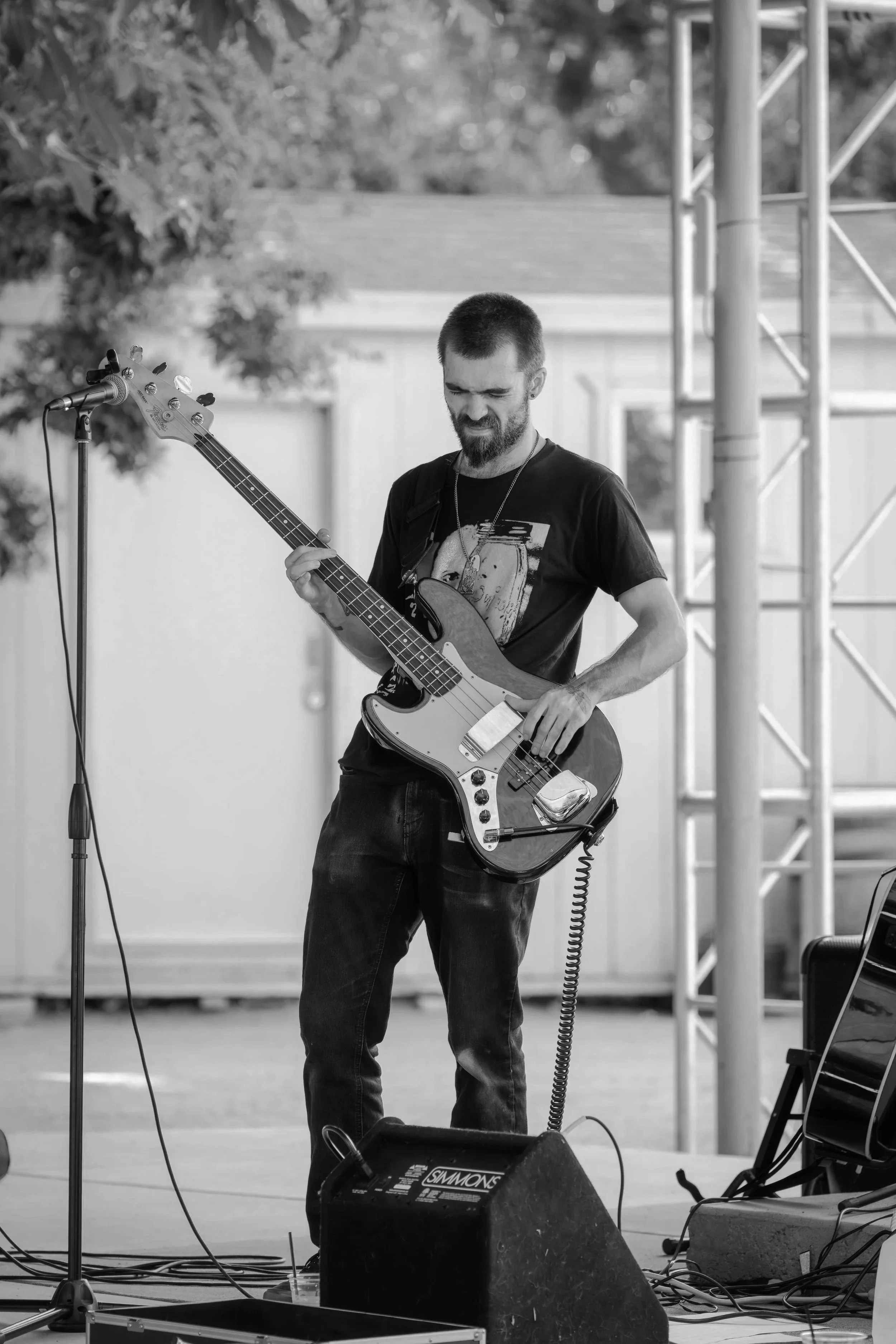 A man with a beard playing an electric bass guitar on an outdoor stage, with a microphone nearby and musical equipment on the ground.