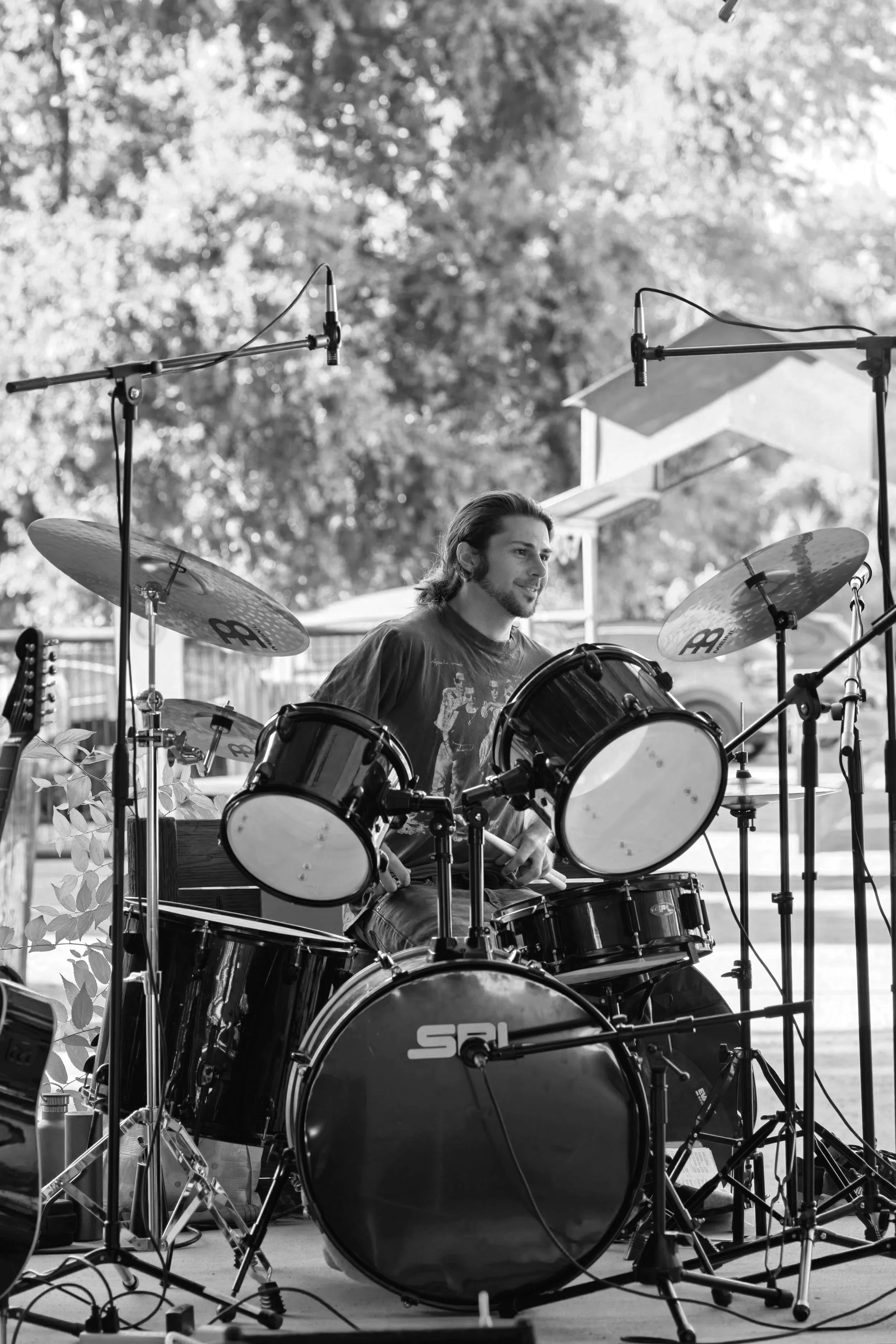 A man playing a drum set outdoors with trees and a car in the background