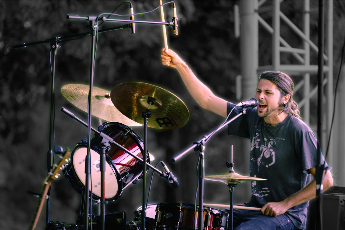 A male drummer performing on stage, singing into a microphone, with drums and cymbals around him, holding drumsticks and raising one in the air.