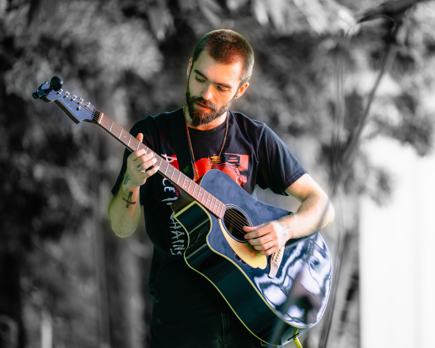 A man with a beard playing an acoustic guitar outdoors, with a blurred black-and-white background of trees.
