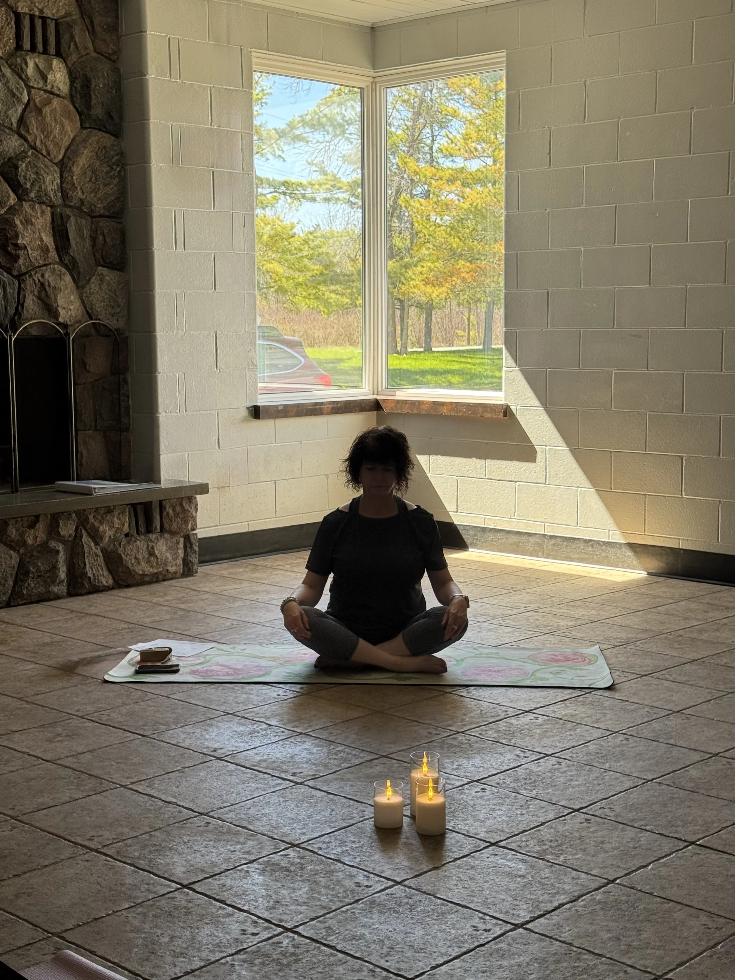 A woman practicing yoga or meditation in a sunlit room with a large window showing trees and grass outside. She sits cross-legged on a mat, with candles placed in front of her.