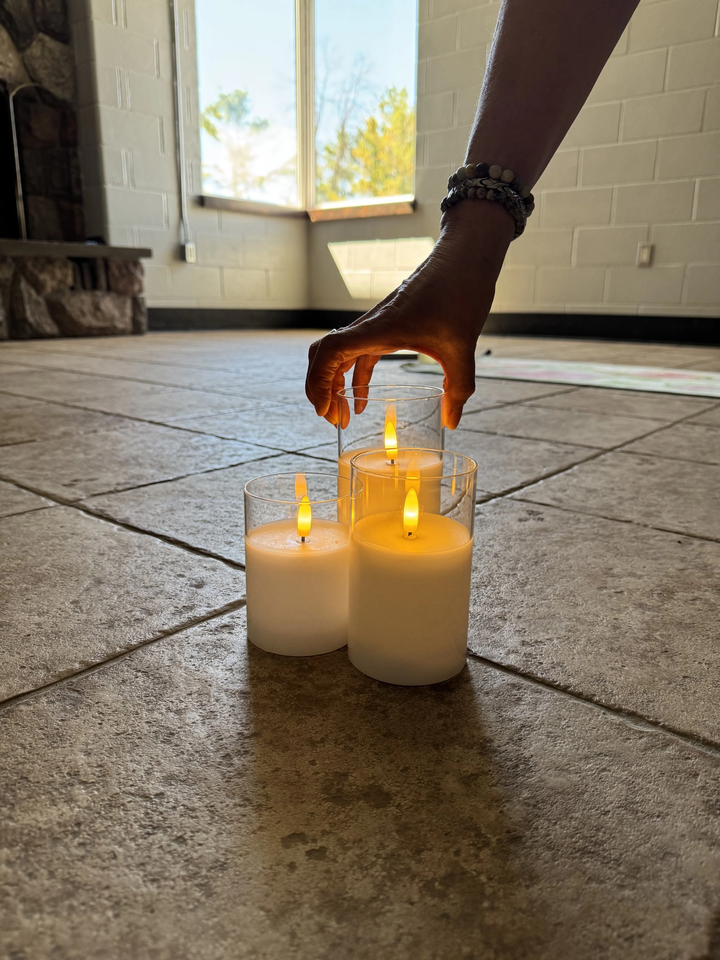 A person placing a lit candle into a glass holder on a tiled floor near an open window with daylight streaming in.
