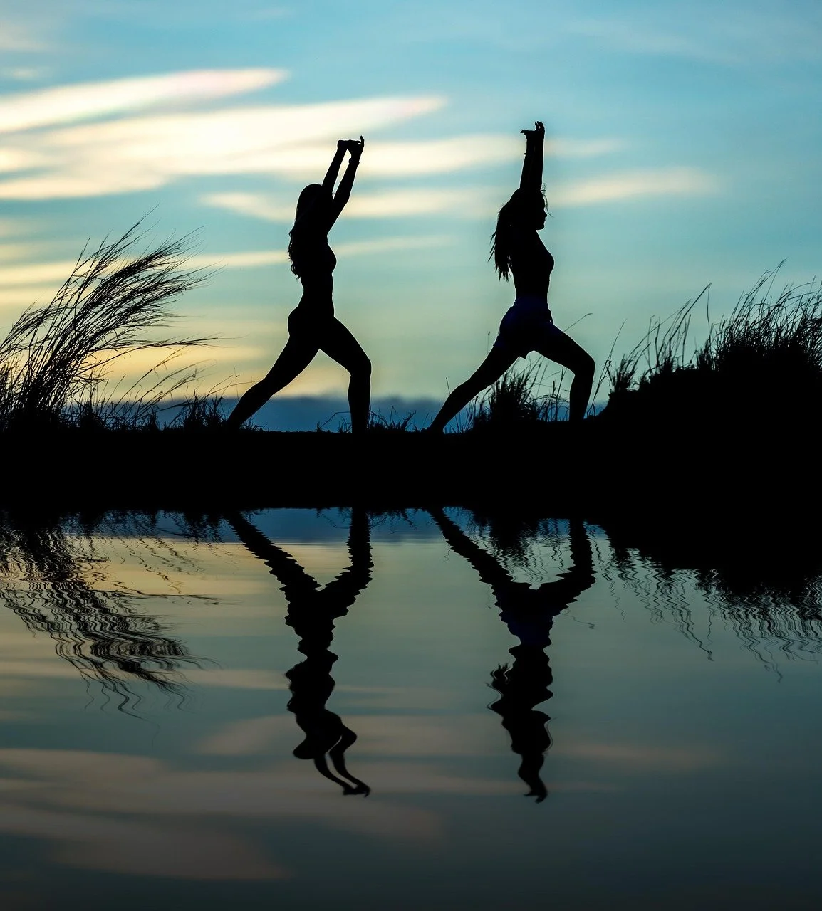 Two women practicing yoga or stretching outdoors during sunset, with their reflections visible in the water below.