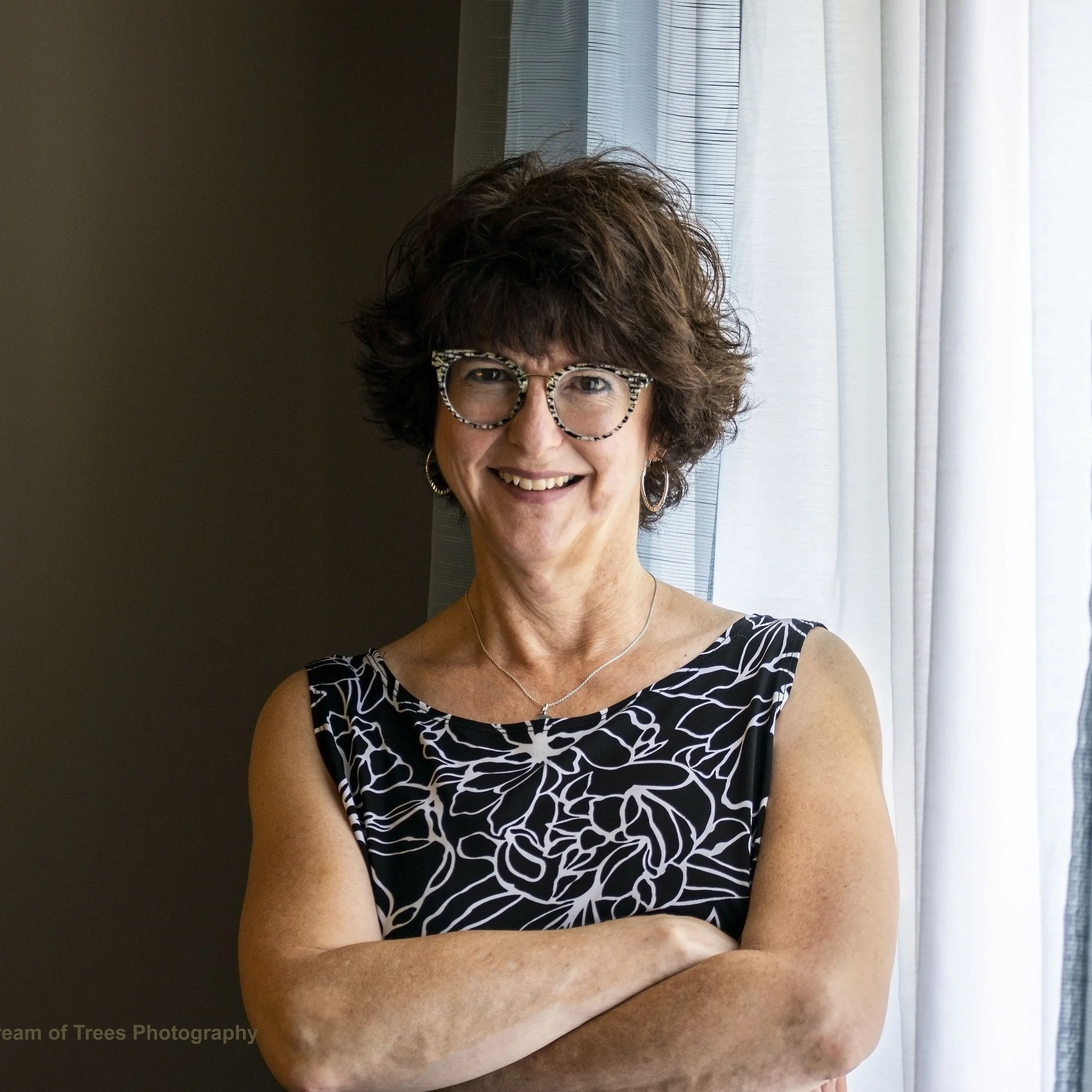 Close-up of a smiling woman with curly brown hair, wearing glasses with a black and white patterned frame, a patterned top, earrings, and a necklace, standing near a window with white curtains.