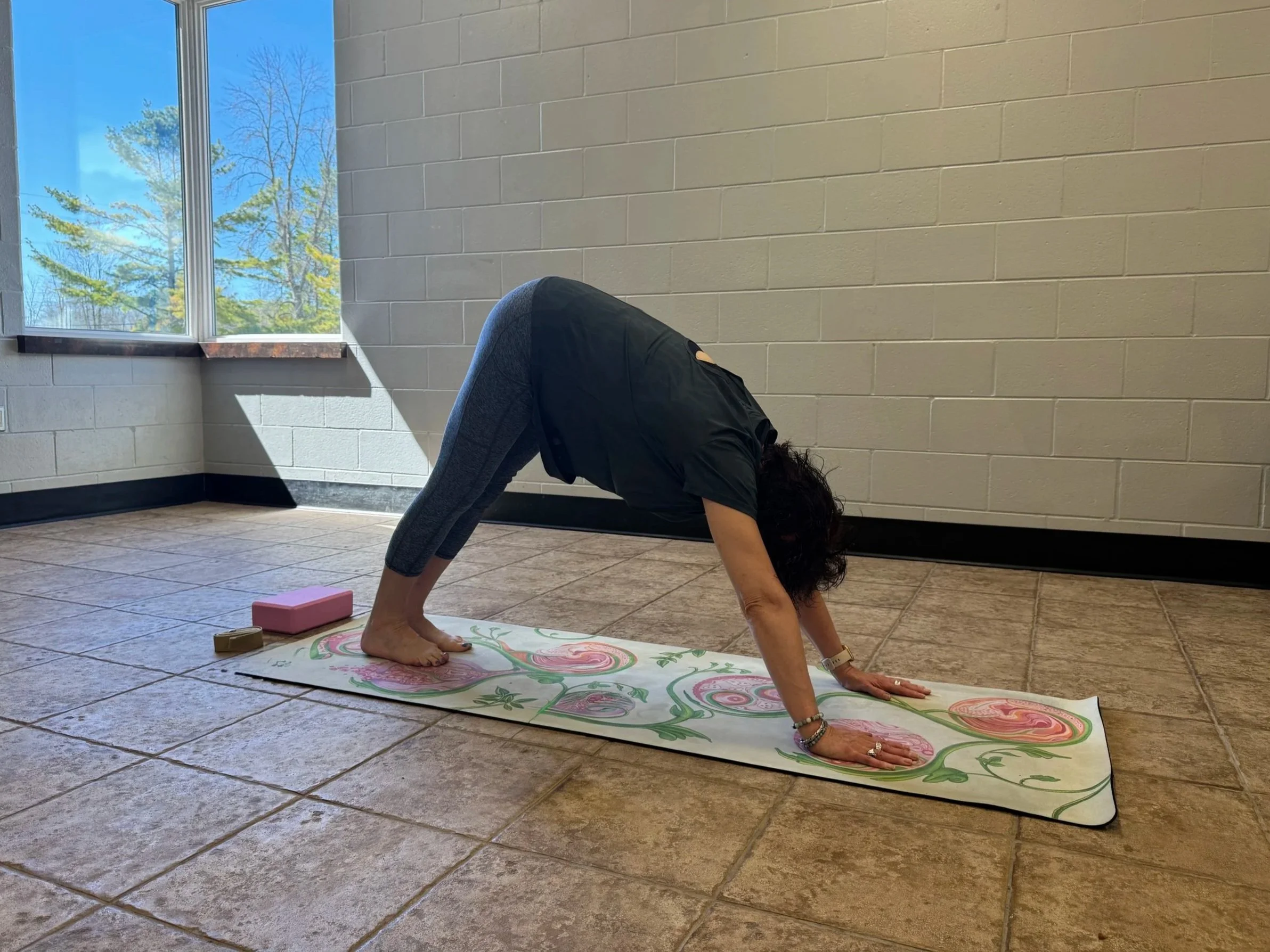 A woman practicing yoga in a room with beige tiled floors and light gray brick walls, stretching in downward dog pose on a colorful yoga mat near a window with tree branches visible outside.