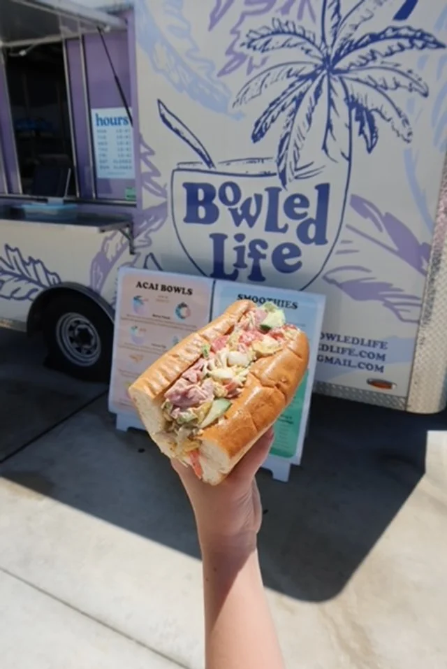 Person holding a hot dog with sliced vegetables and sauce in front of a food truck with 'BOWLED LIFE' signage and a palm tree illustration.