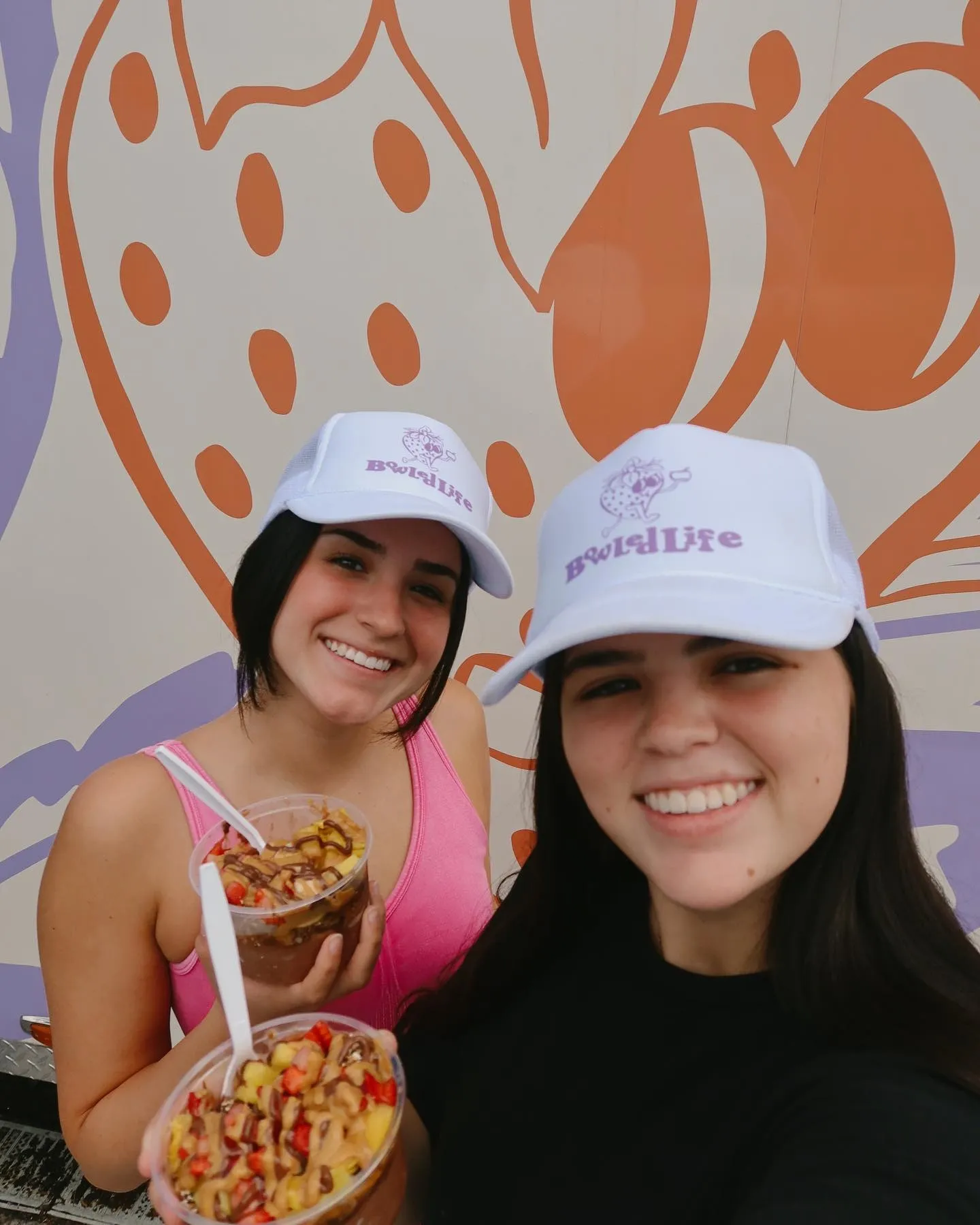 Two women smiling in front of a colorful wall, wearing white hats with purple lettering, holding bowls of ice cream topped with fruit and chocolate drizzle.