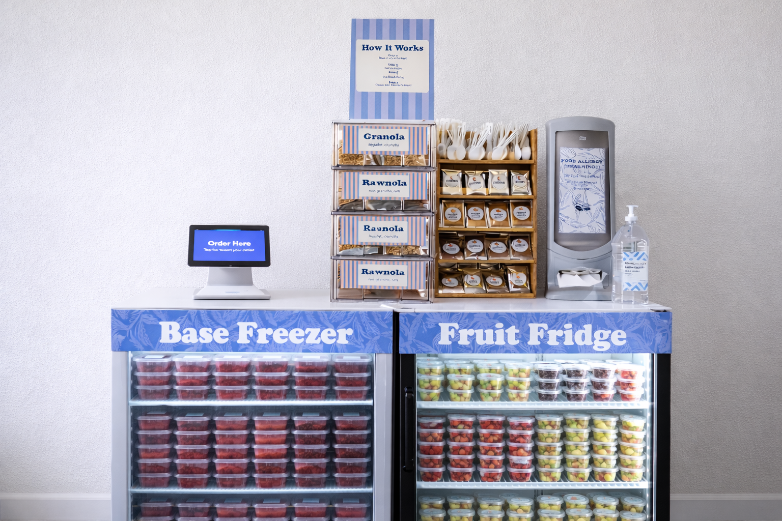 A display of pre-packaged fruits in a refrigerated case labeled 'Base Freezer' and 'Fruit Fridge'. The freezer contains rows of strawberries, and the fridge contains containers of various chopped fruits. There is a small digital ordering kiosk, snack boxes labeled 'Granola' and 'Rawnola', a hand sanitizer bottle, and a machine with utensils on top.