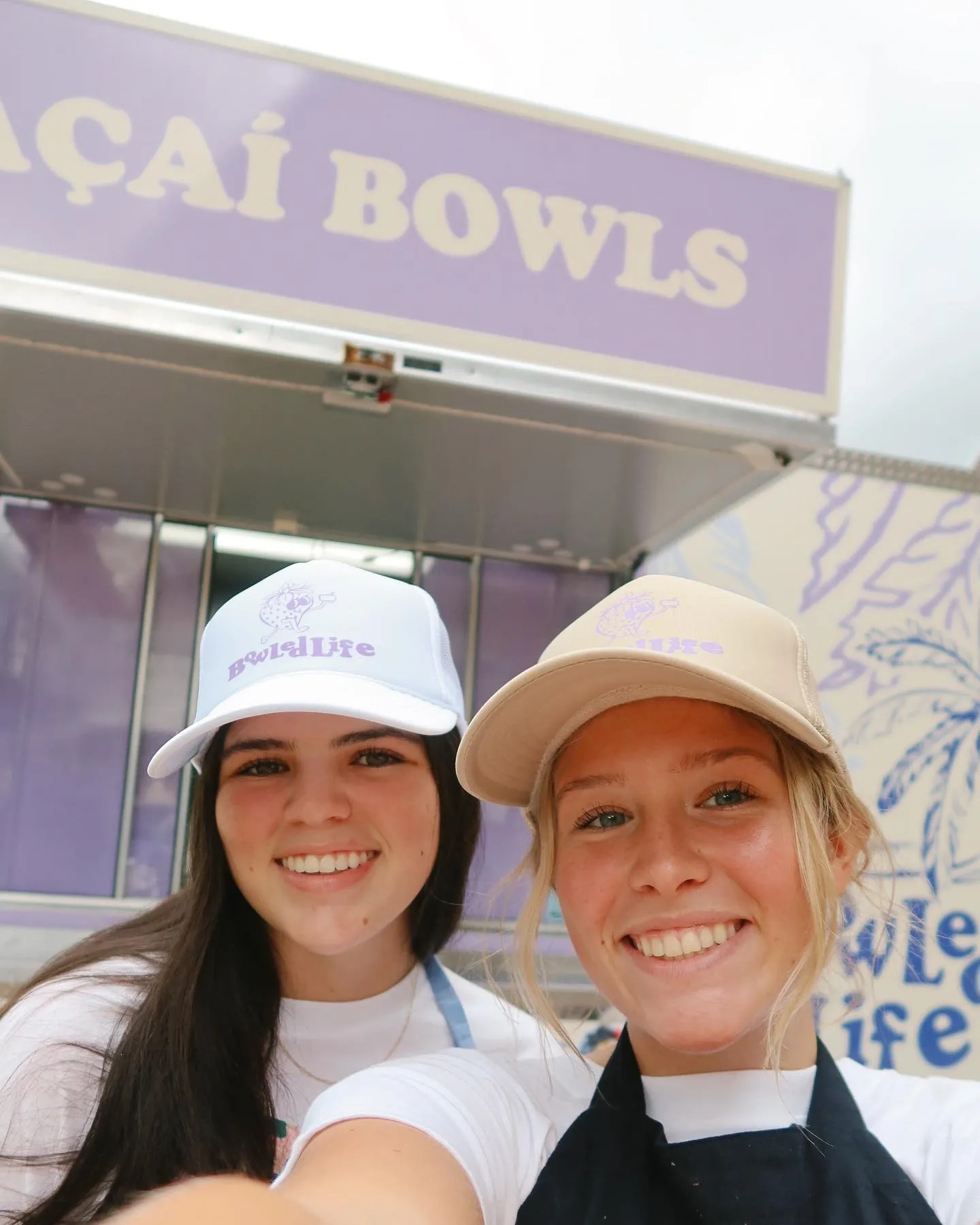 Two young women smiling and taking a selfie in front of a purple and white booth that says 'Bowl Life'. Both women are wearing white caps with the 'Bowl Life' logo and casual clothing.
