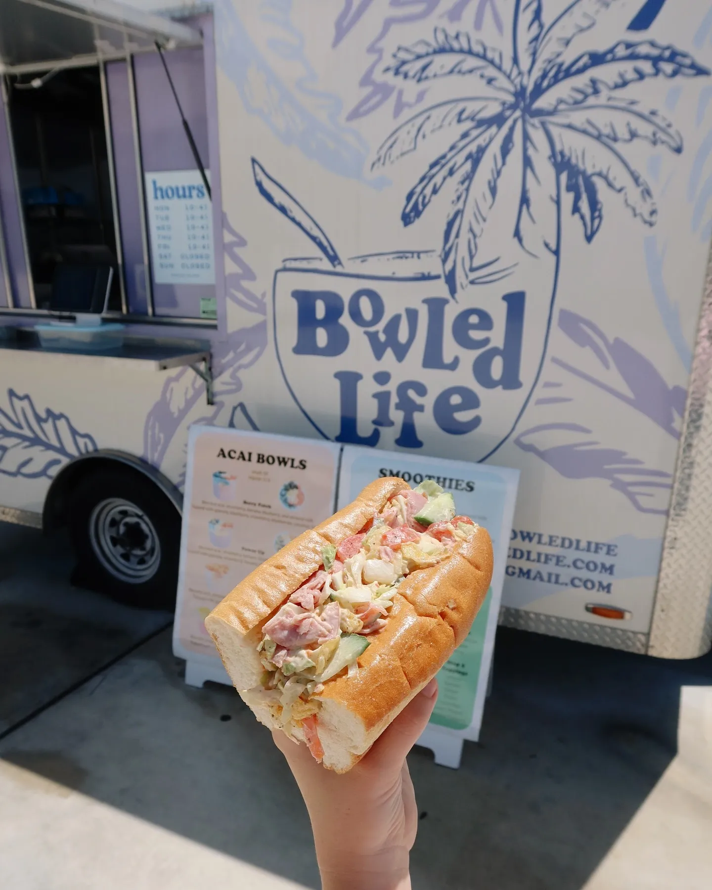 A hand holding a sandwich with chopped vegetables and possibly chicken, in front of a food truck with a large sign that reads 'Bowl'd Life' and a drawing of a coconut drink with a straw and a palm tree.