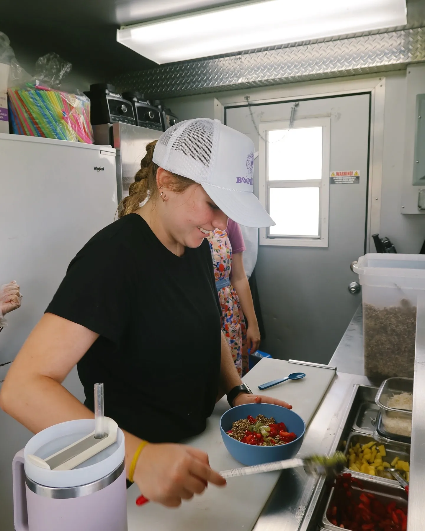 A smiling woman wearing a white baseball cap and black t-shirt preparing a colorful bowl of food in a kitchen.