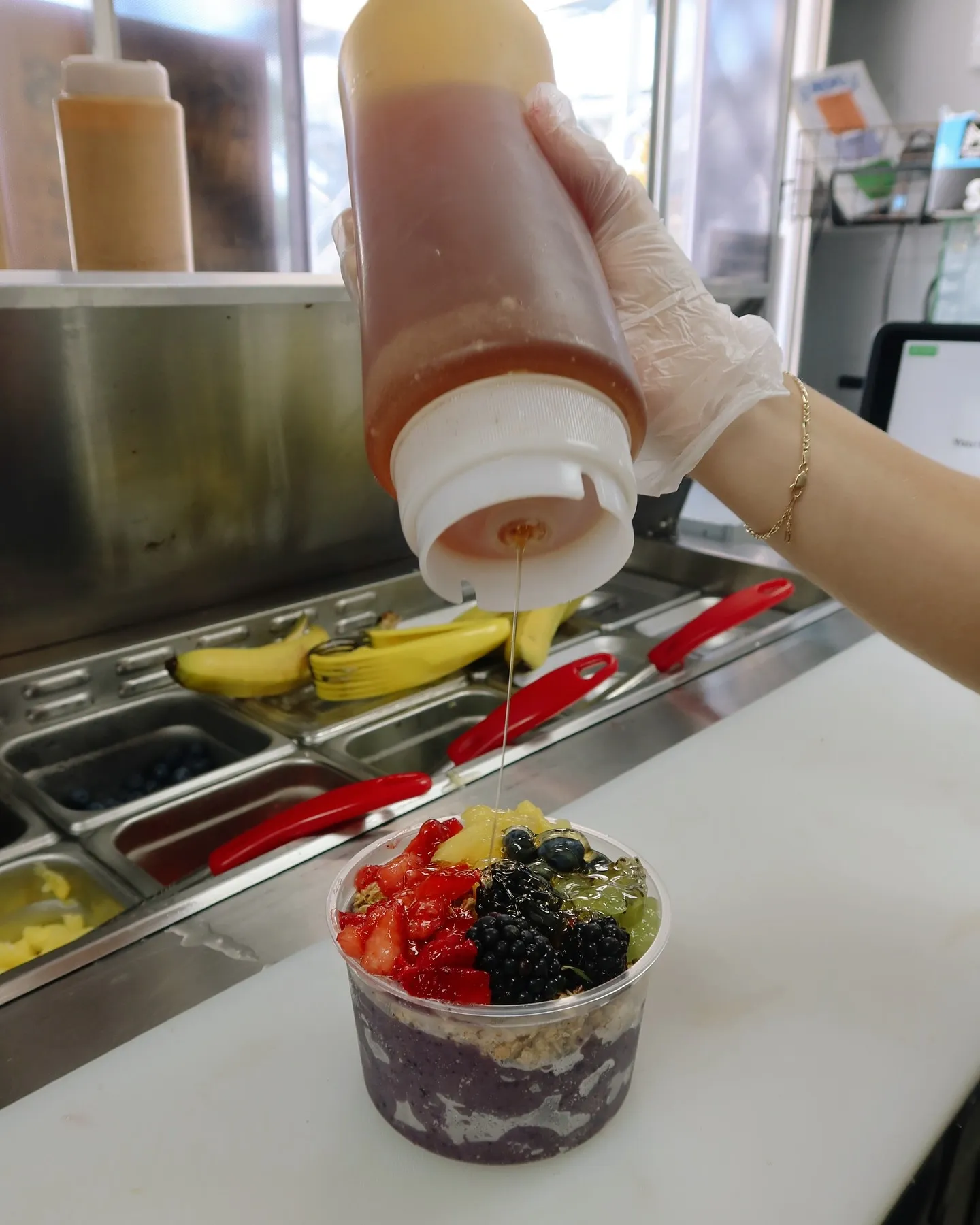 A person wearing a glove is pouring chocolate syrup from a squeeze bottle onto a bowl of mixed fruit and granola. The bowl contains strawberries, blackberries, kiwi, and blueberries.