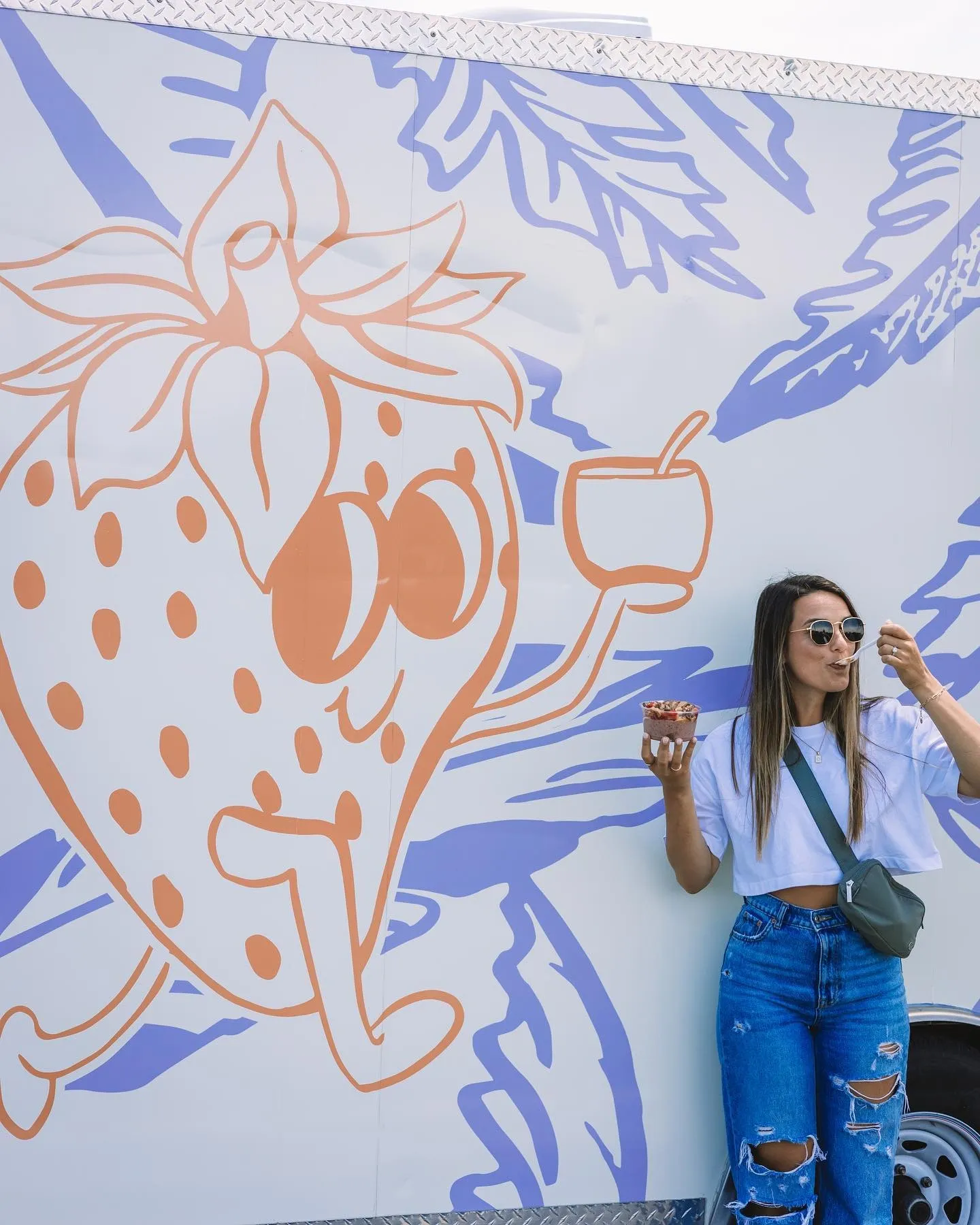 A woman with long hair wearing sunglasses, a white crop top, and ripped jeans, standing next to a large white trailer with a colorful mural of a strawberry, leaves, and a bowl of strawberries. She is holding a bowl of strawberries and eating a strawb