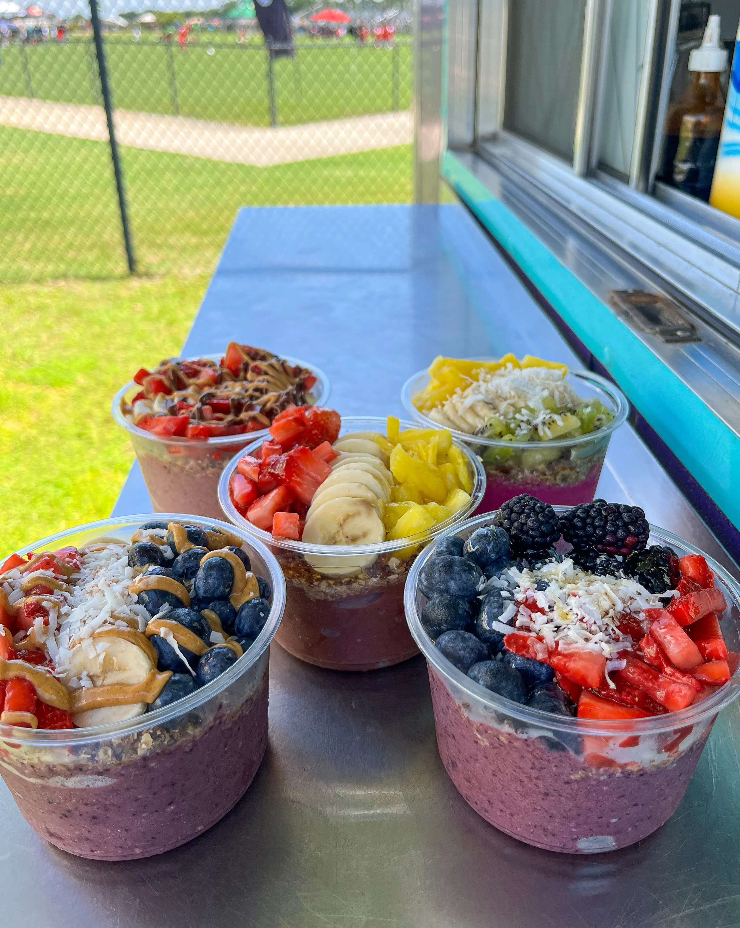 Six cups of colorful smoothie bowls topped with fresh fruit, nuts, and sauces, placed on a metal counter outdoors.