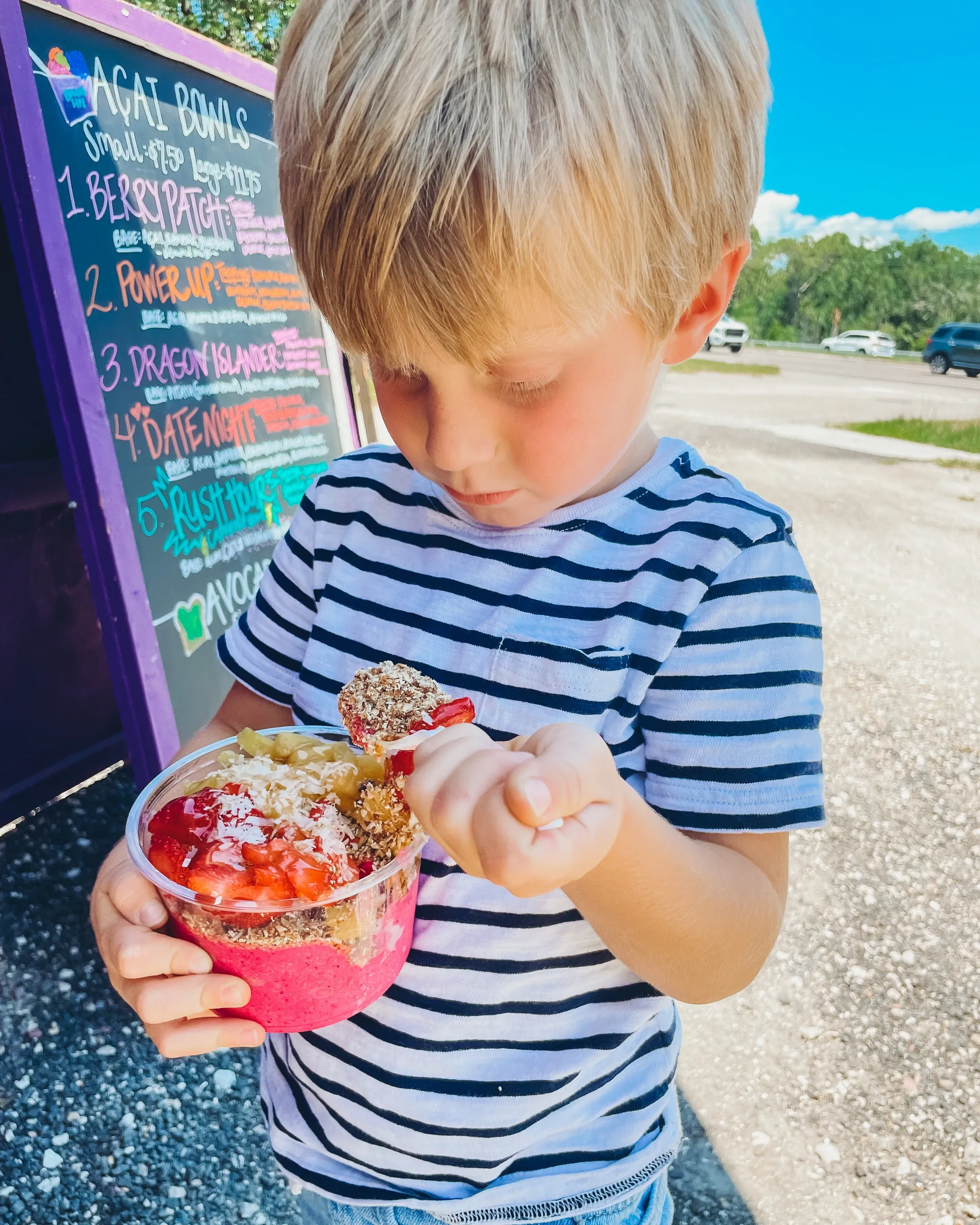 A young boy in a striped shirt holding a cup of frozen yogurt with strawberries and toppings, standing outside near a colorful menu board on a bright, sunny day.