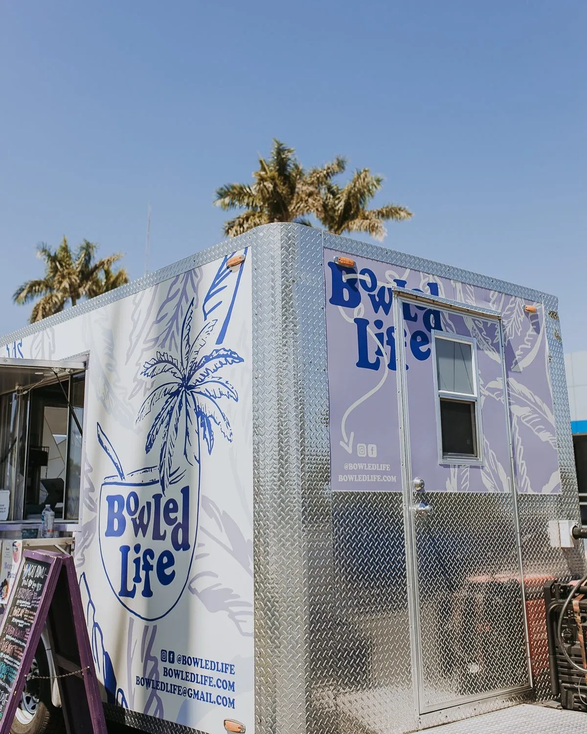 A food truck with a white and silver exterior, featuring the logo 'Bowled Life' with a palm tree illustration and contact information.