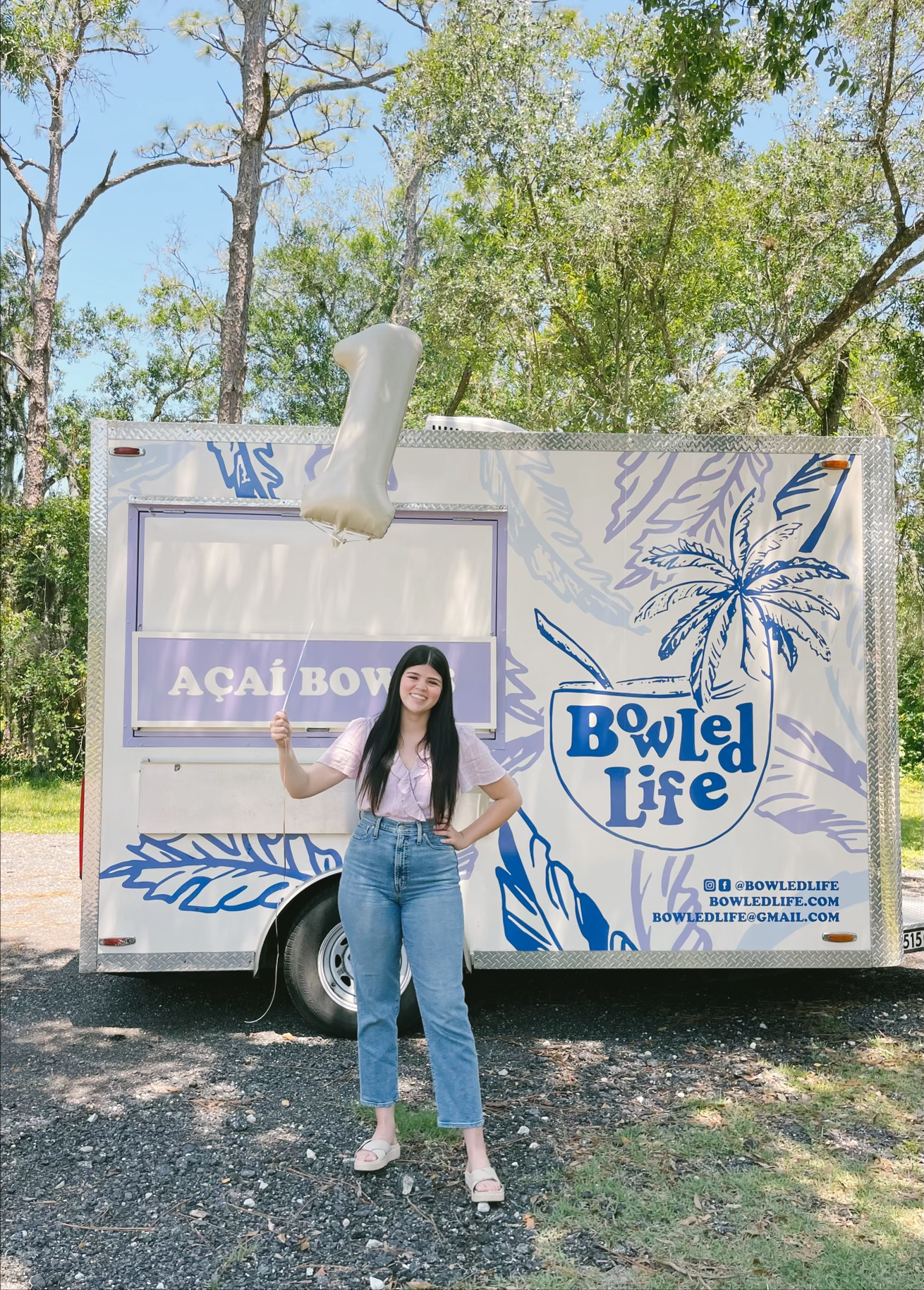 Young woman holding a balloon in front of a food truck with 'Bowl Life' logo and tropical design, surrounded by trees and gravel ground.