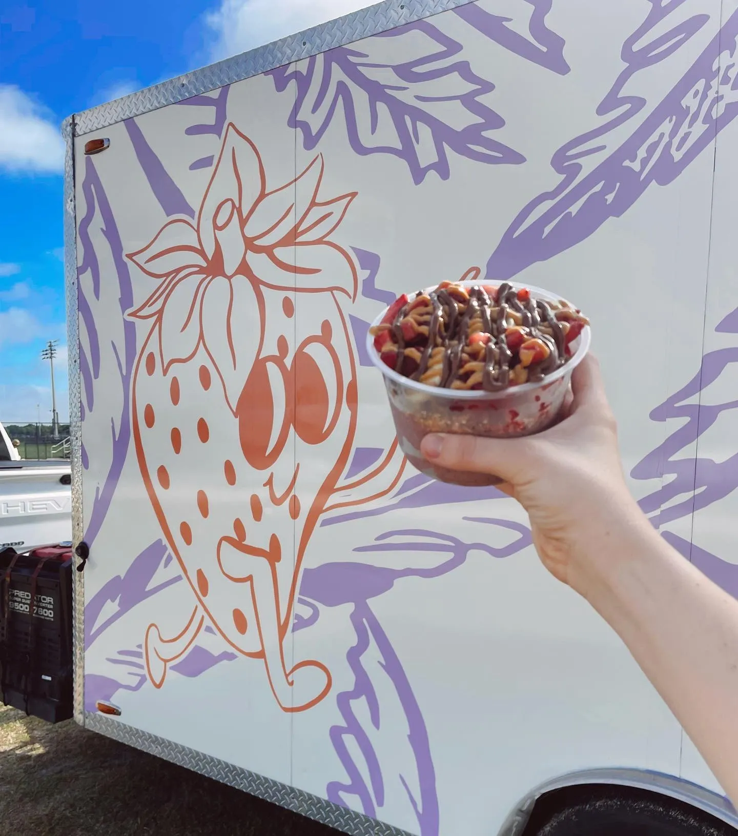 Hand holding a bowl of ice cream topped with chocolate and caramel drizzle in front of a food truck with a strawberry illustration.
