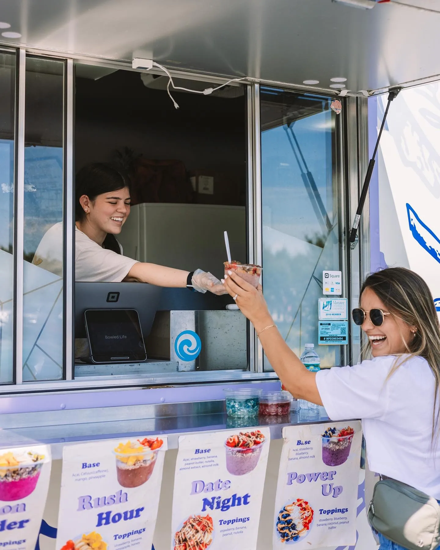 A woman with sunglasses receives a strawberry smoothie from a smiling female vendor at a food truck. The truck displays colorful signs for different smoothie flavors.
