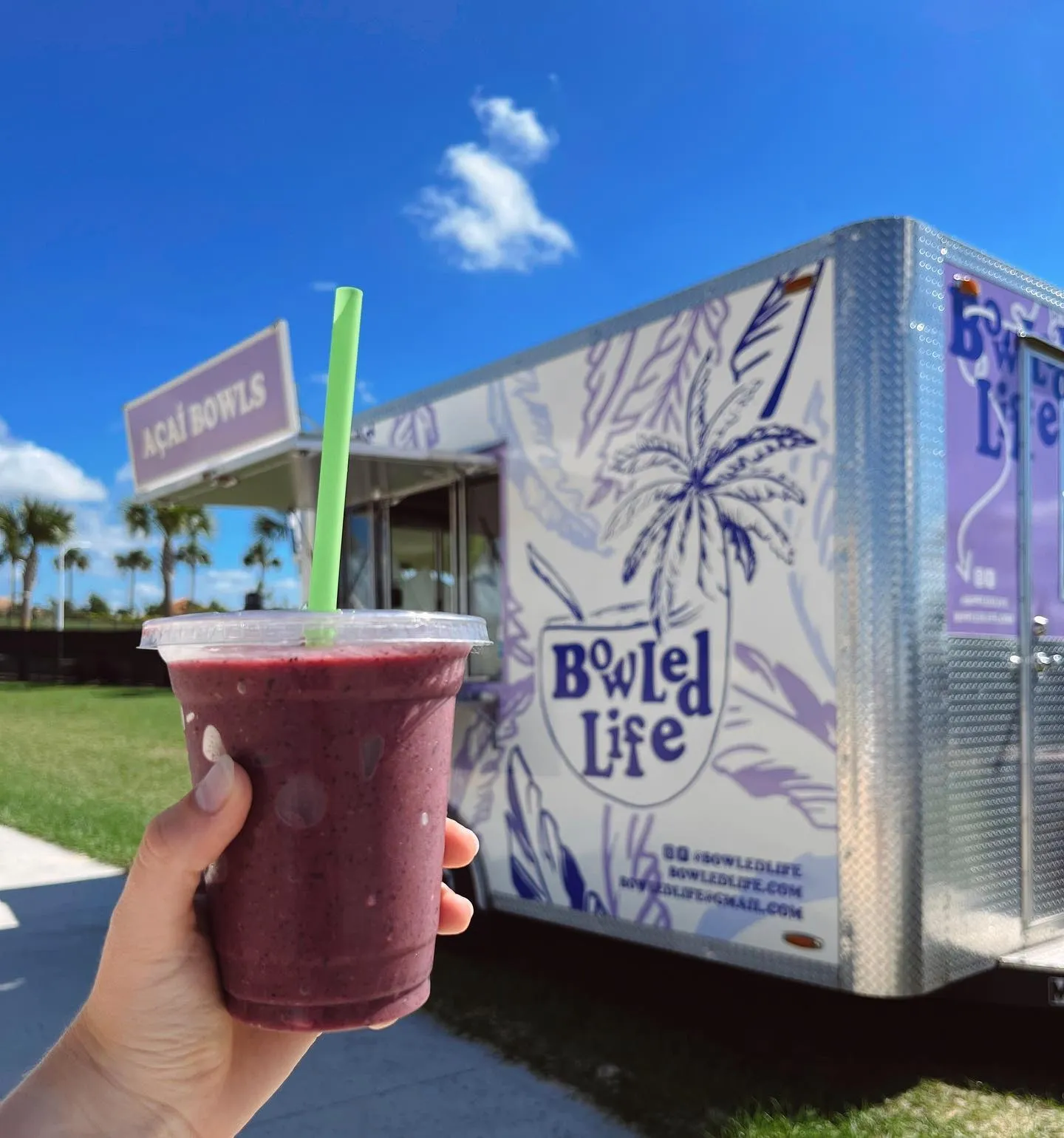 A person holding a purple berry smoothie with a green straw in front of a food truck called 'Bowl'd Life' which has a purple palm tree logo and a white and purple design.