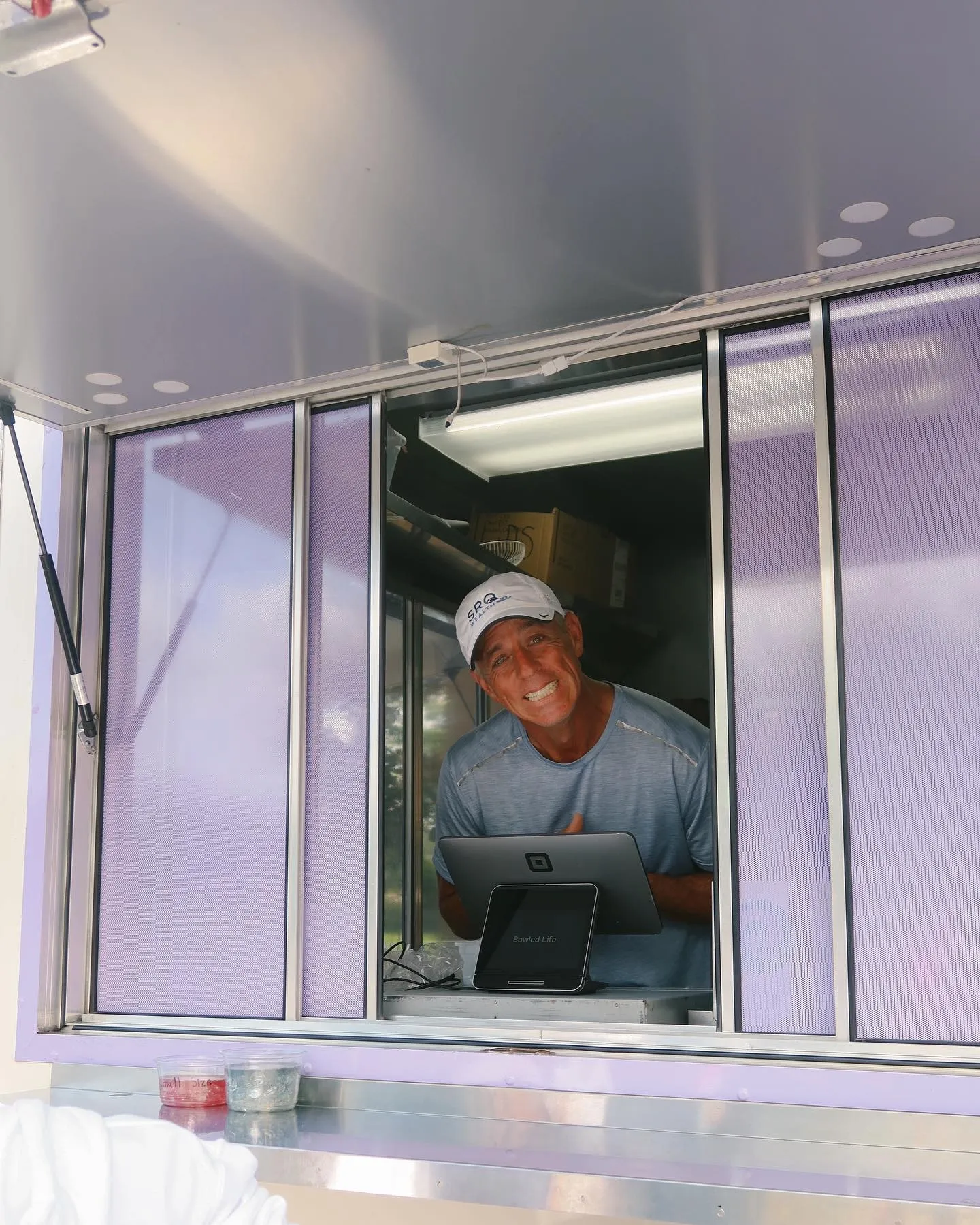 A man smiling and leaning forward through a food truck window, wearing a white cap and a light blue shirt. There are containers with drinks in front of him and a tablet device on the counter.