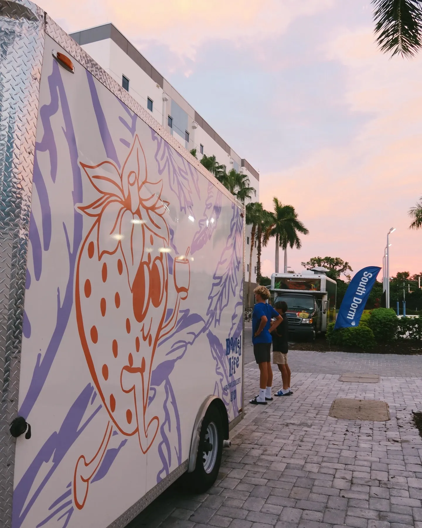 A food truck with a large strawberry illustration on its side, parked on a brick-paved area during sunset, with two young boys standing nearby and a South Dade banner in the background.