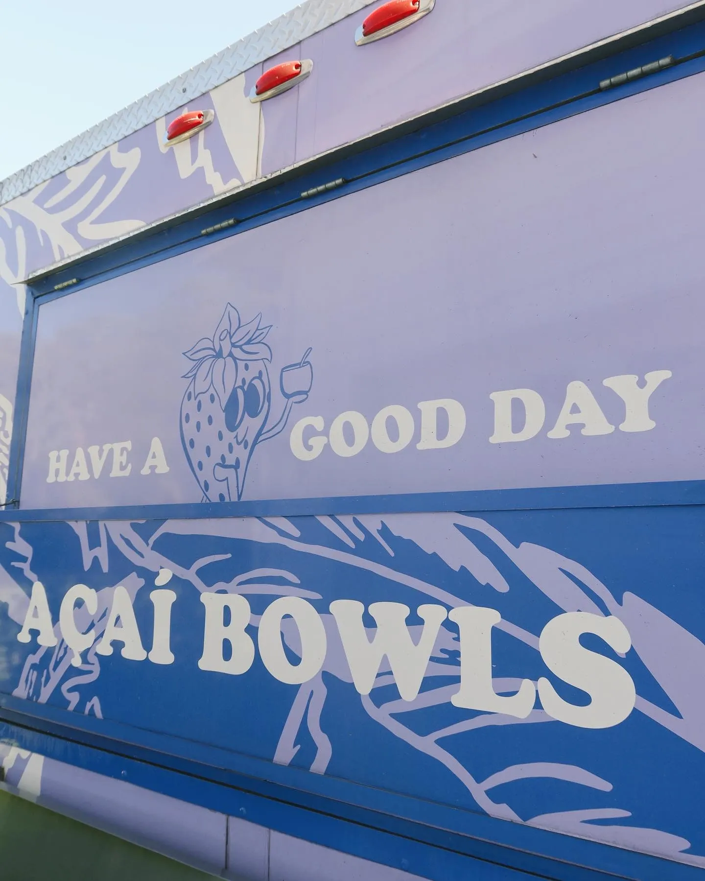 Sign with a cartoon strawberry character holding a drink that says 'Have a good day' and 'Acaí Bowls' on a blue background.