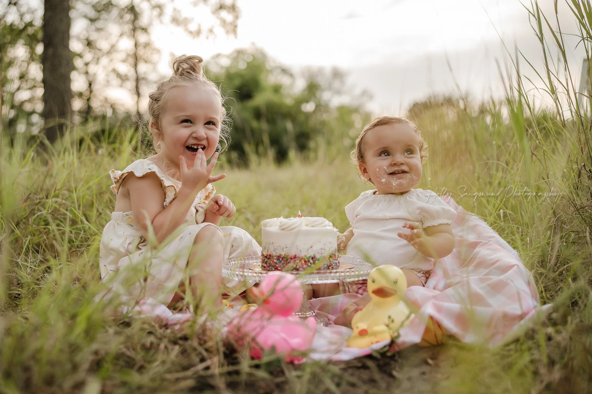 Sisters eating cake during a first birthday smash cake session outside in the grass in Lexington, Texas