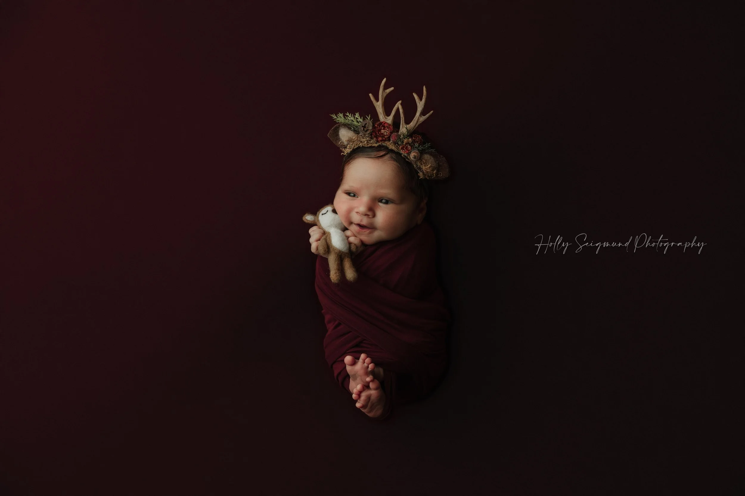 Baby girl on a maroon backdrop and wrapped in a maroon wrap, holding a felt baby deer with a floral deer antler crown. In-studio newborn session in Lexington, Texas