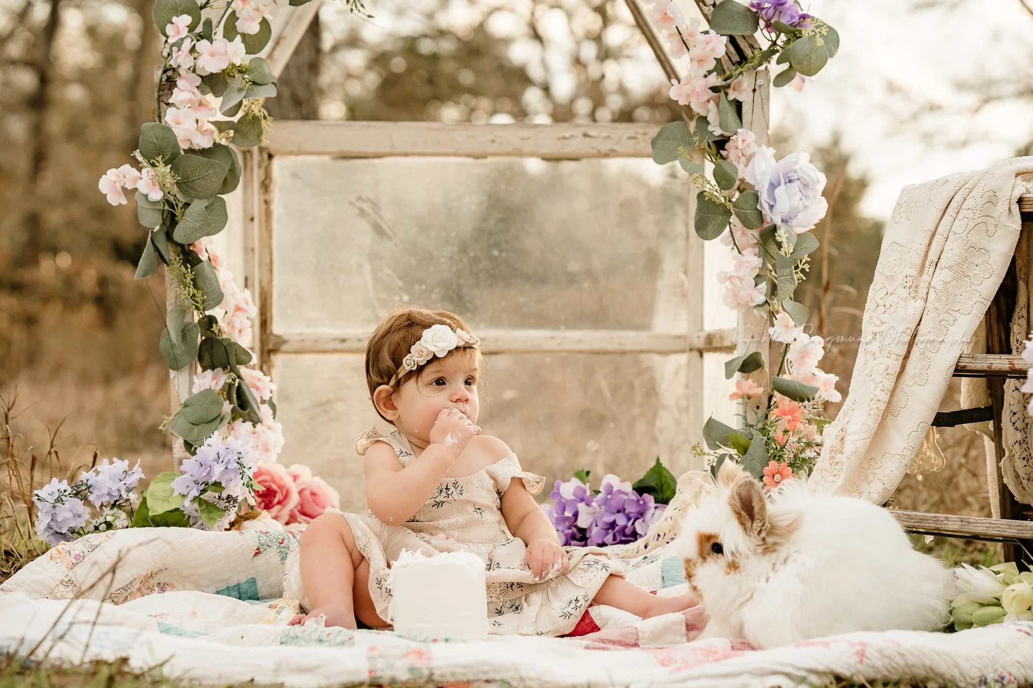 One year old girl eating cake during a smash cake session in front of a flower covered mini greenhouse in Central Texas
