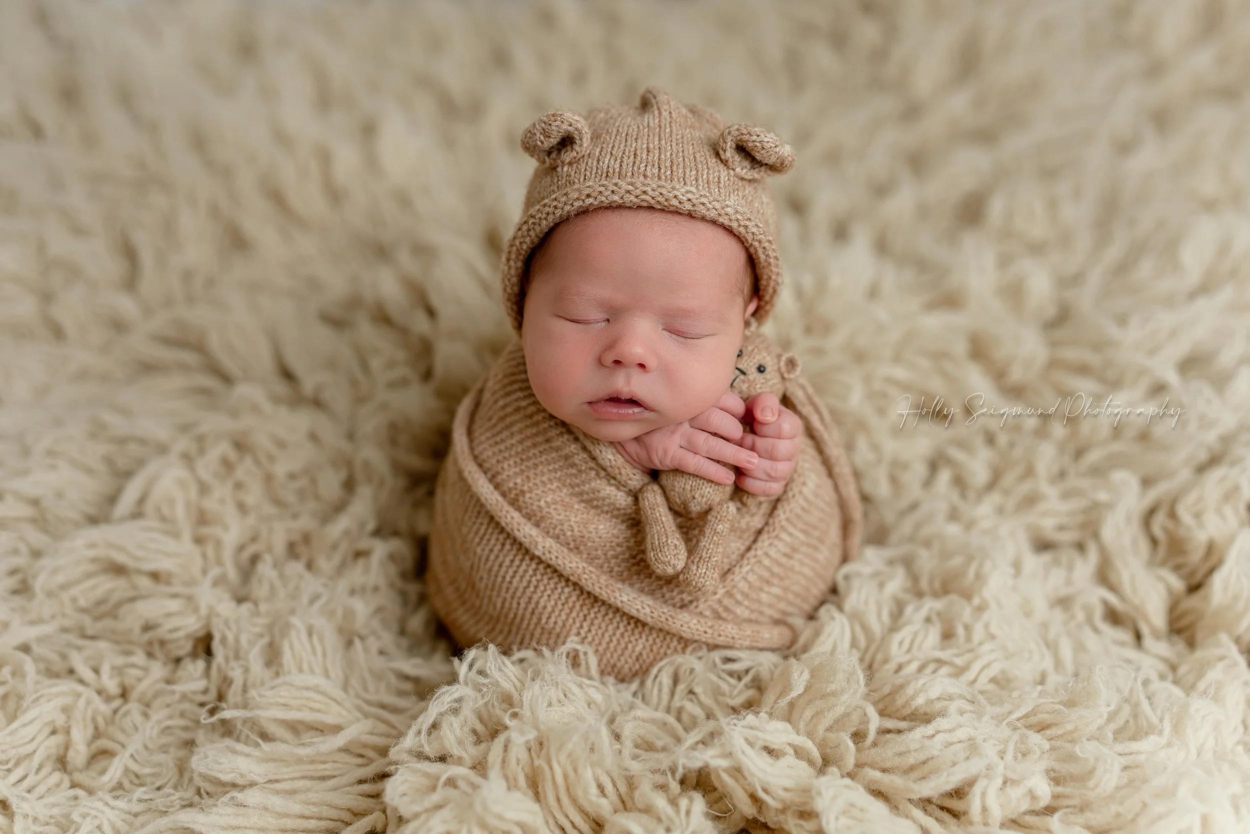 Newborn baby boy wrapped in a neutral tan swaddle with a bear bonnet, holding a tan teddy bear on a tan fluffy backdrop in Blue, Texas