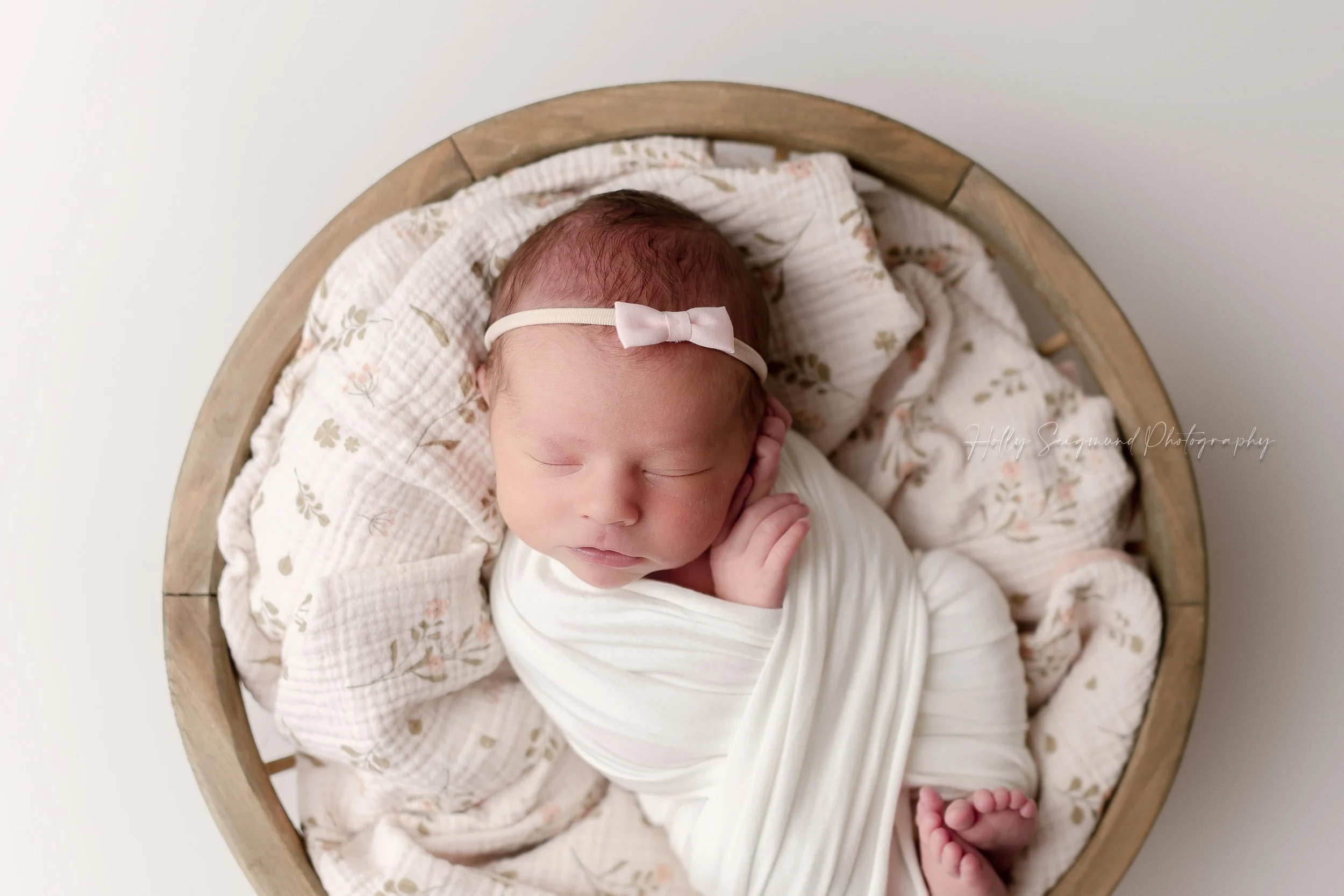 Newborn baby girl wrapped in white with a tiny pink bow, posed in a round wooden bowl with hands and toes peeking out of the wrap during a studio newborn session serving Elgin, Texas