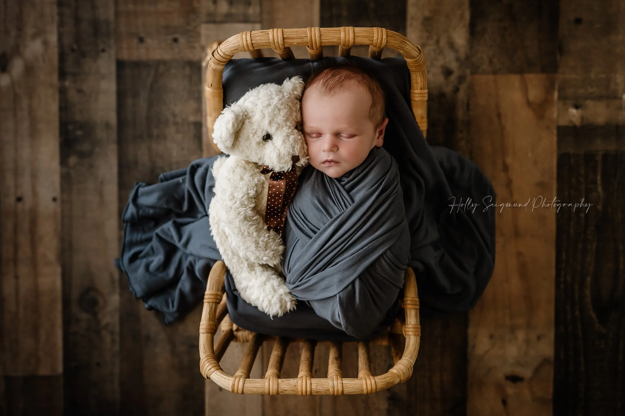 Newborn baby boy wrapped in blue with his teddy bear on a wood bed on a wood floor in a Lee County Texas Studio