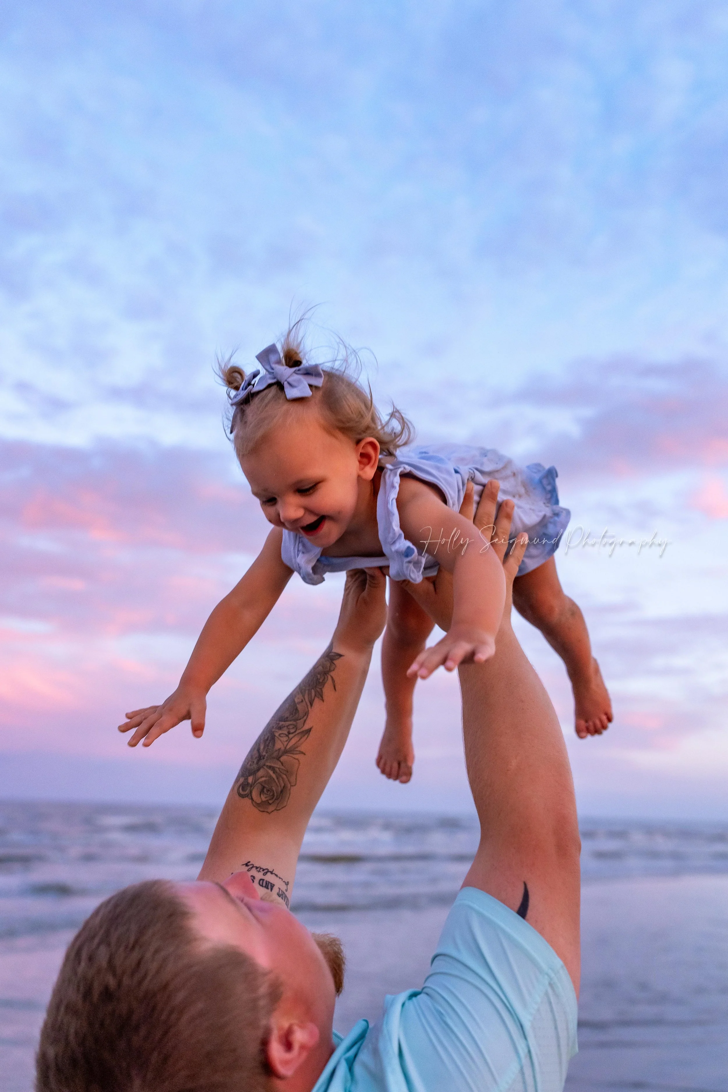 Dad playing with daughter with the pink and purple Texas sunset in the background during Beach Mini Session in Port Aransas