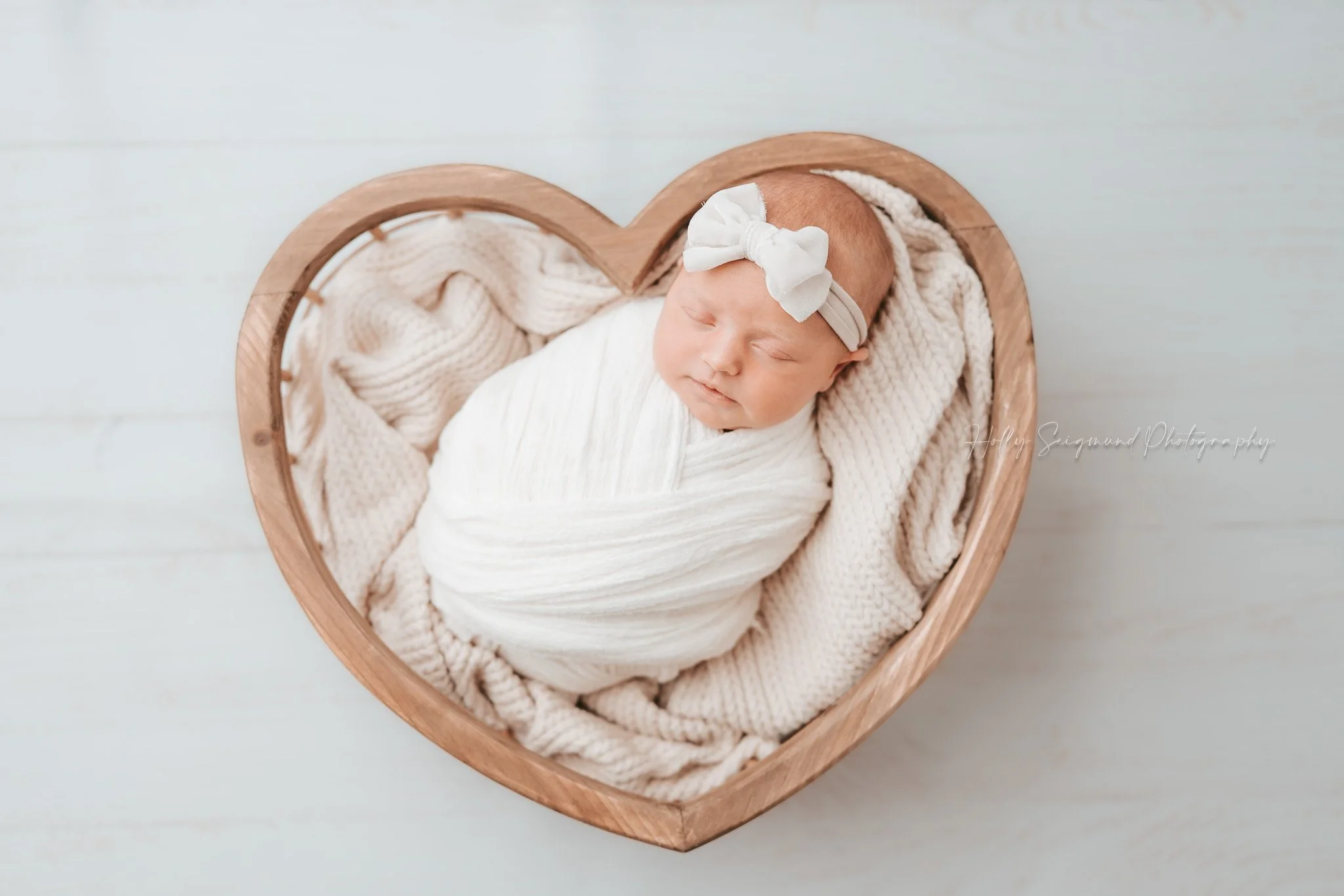 Newborn from Central Texas in wooden heart bowl with white wrap and bow on a white floor