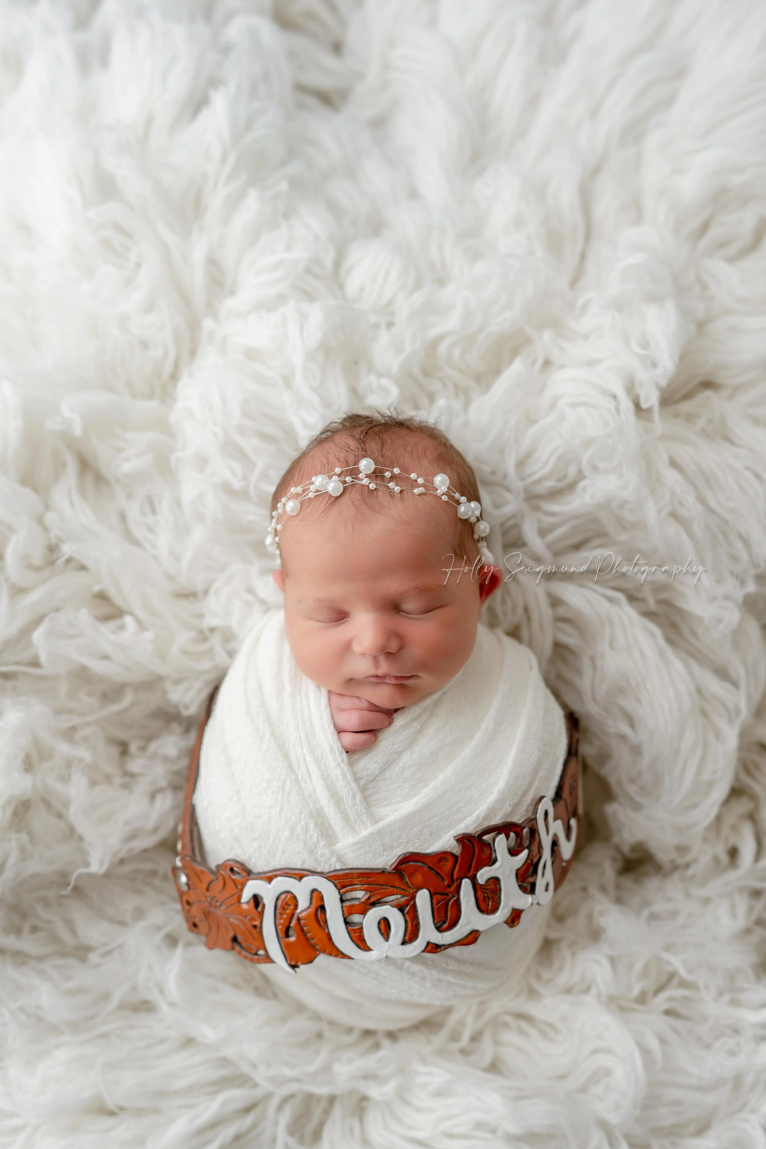 Newborn baby girl wrapped in white on a white fluffy backdrop, pearl headband and mom's personalized leather wedding dress belt during a studio session in Blue, Texas