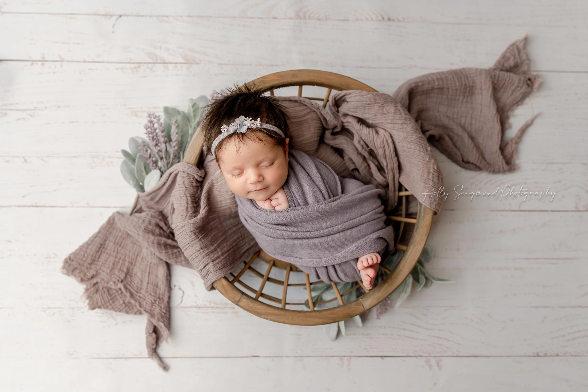 Wrapped newborn baby girl in purple, posed in a round wooden bowl in a studio newborn session in Lee County Texas