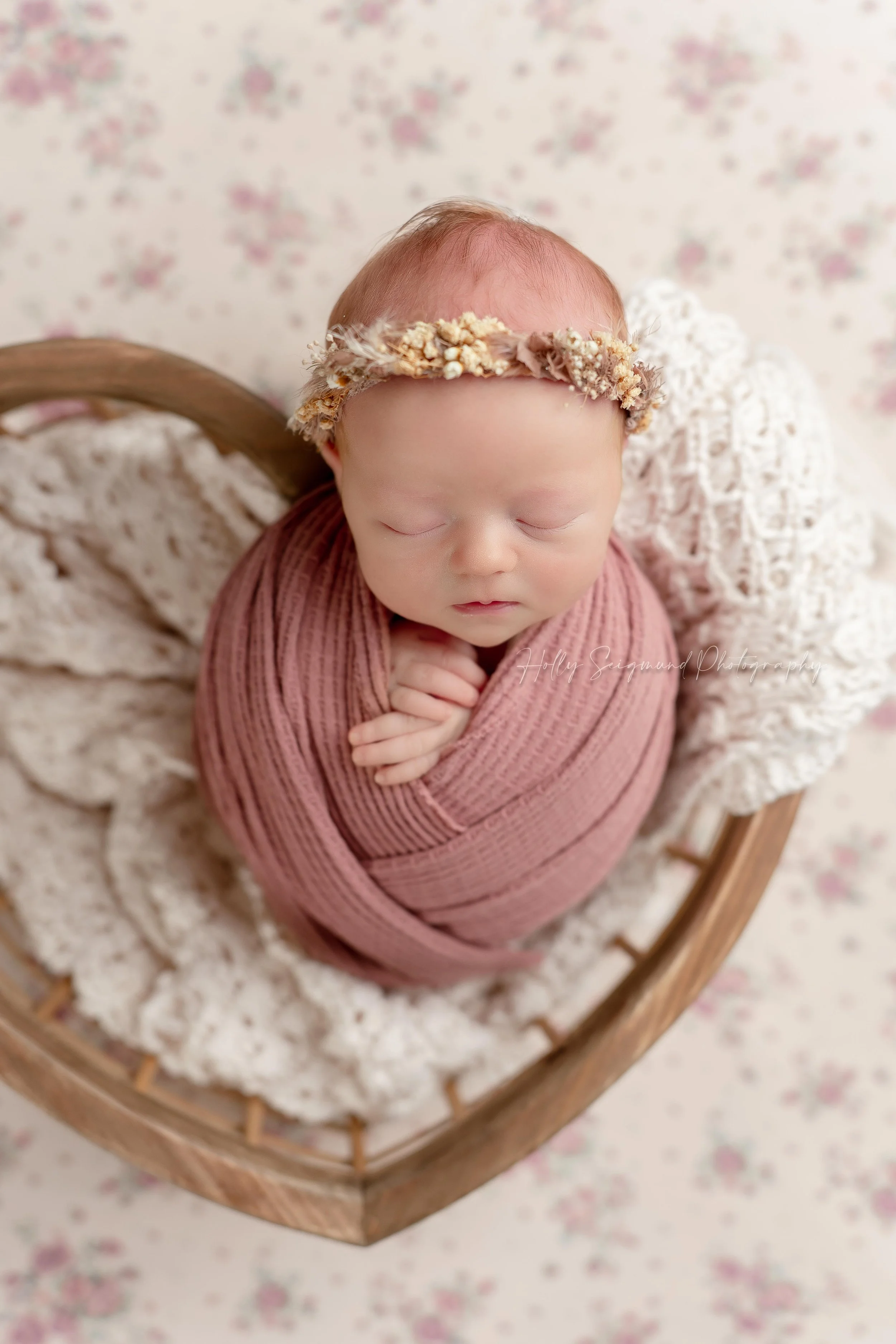 Baby girl wrapped in a pink wrap, posed in a wooden heart crate on a floral pink and white backdrop in Bastrop County