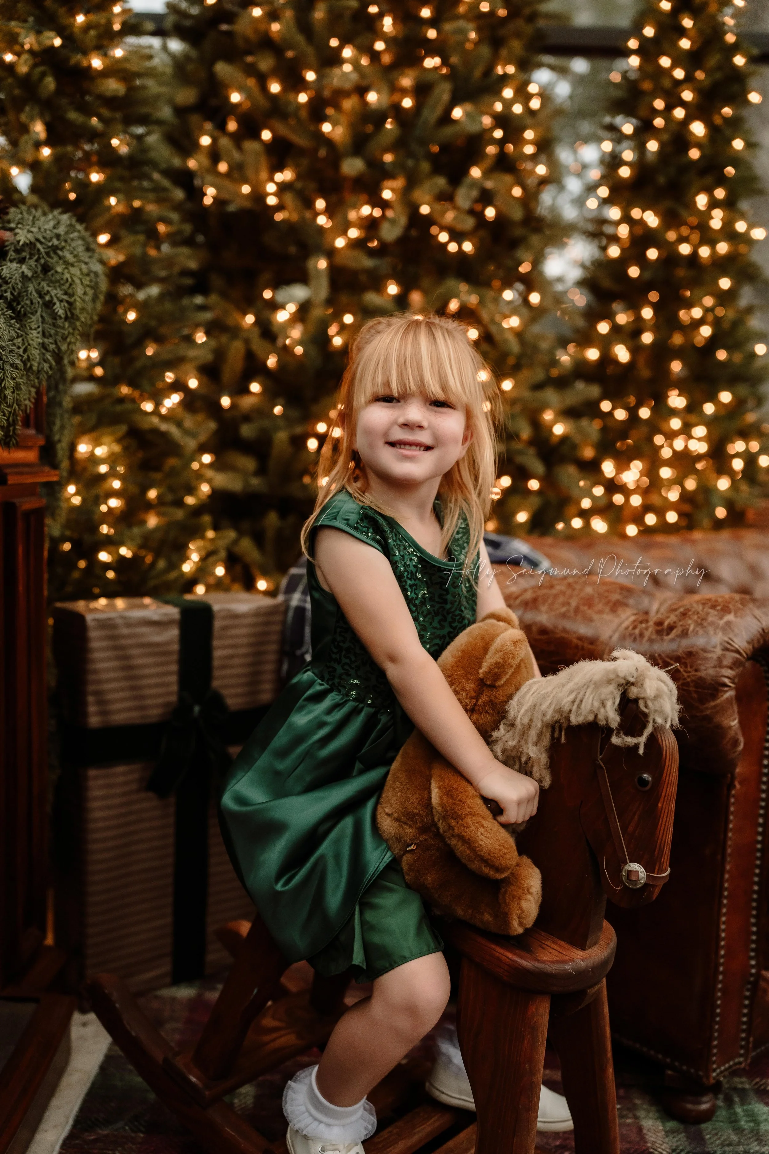 Smiling young girl in an emerald green Christmas dress sitting on a wooden rocking horse, surrounded by warmly lit Christmas trees during a holiday mini session with Holly Seigmund Photography in Blue, Texas.