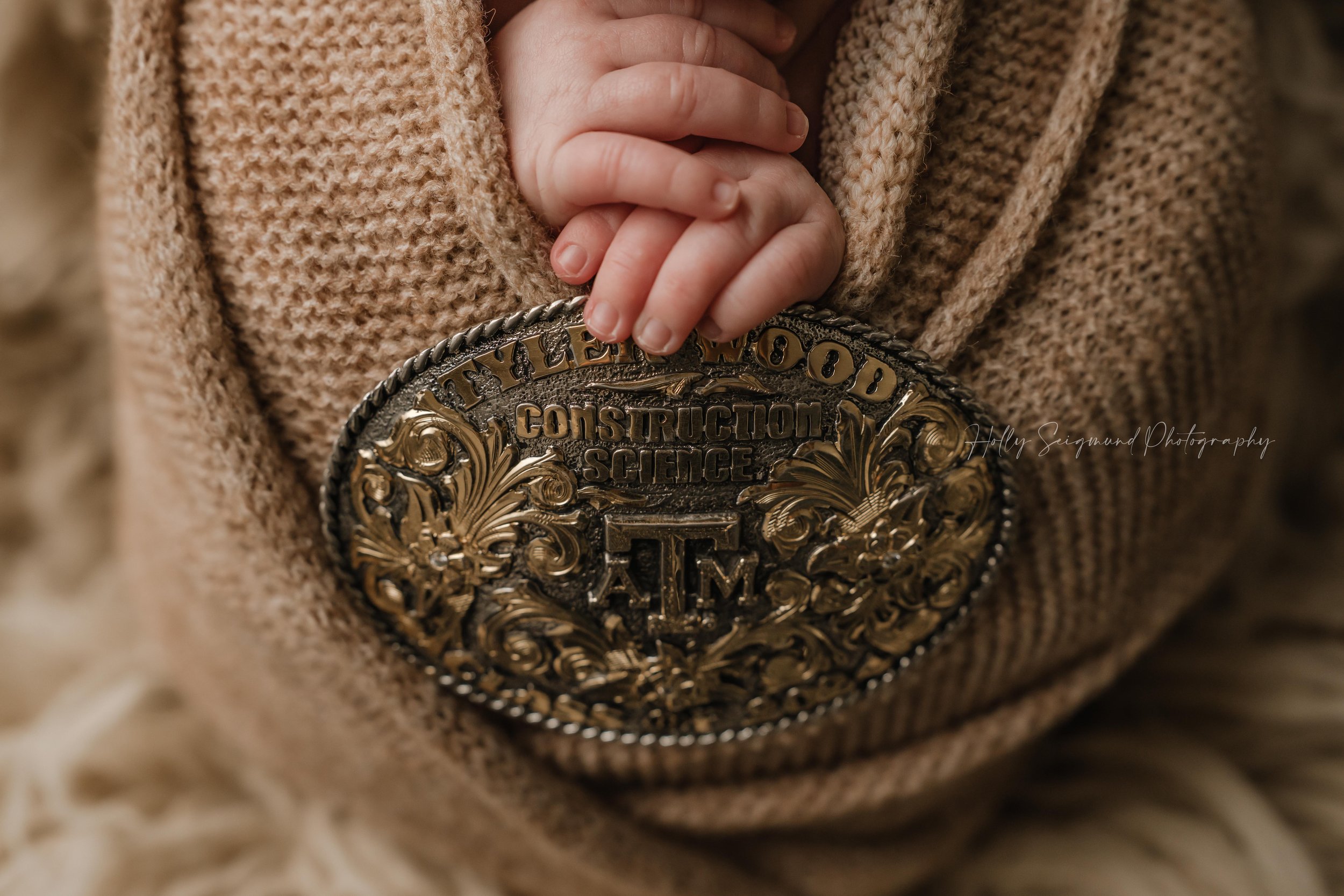 Close up detail shot of a wrapped baby boy with a tan knit wrap, holding his dad's A&M belt buckle during a in-home studio newborn session in the Bastrop County area