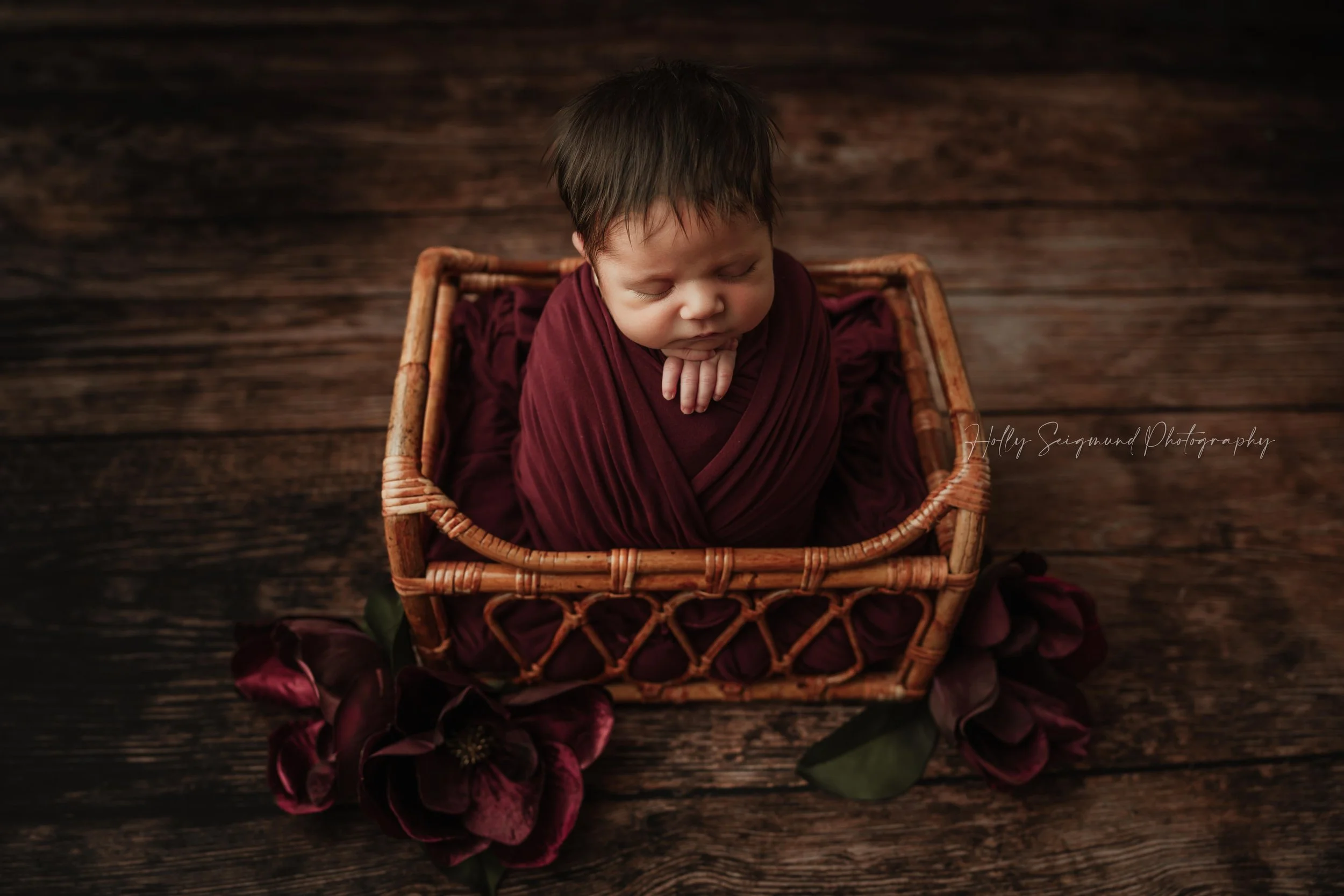 Newborn baby with maroon wrap, posed in a wooden baby bed with maroon flowers on a wooden floor backdrop during a studio newborn session in Blue, Texas