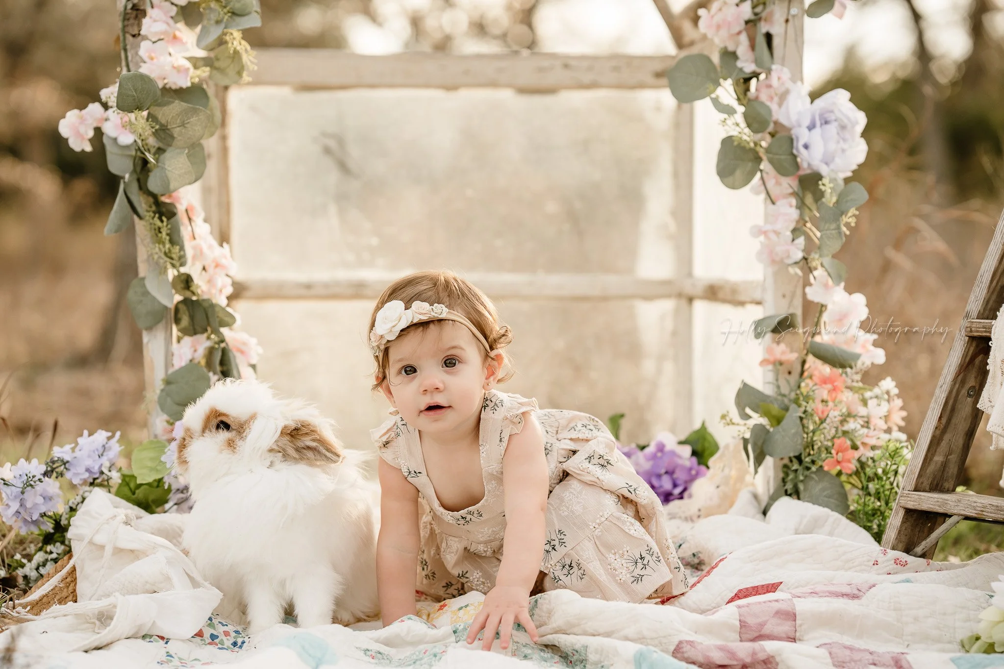 One year old and bunny in front of a mini greenhouse with spring flowers on a quilt for first birthday photos in Lee County Texas