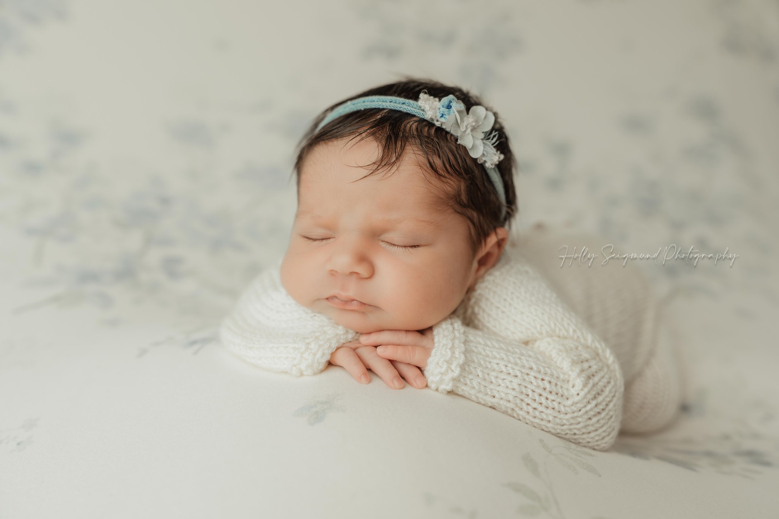 Posed newborn baby girl with head on hands, on a cream and blue floral backdrop with blue headband and white knit outfit during a studio newborn session serving Elgin, Texas