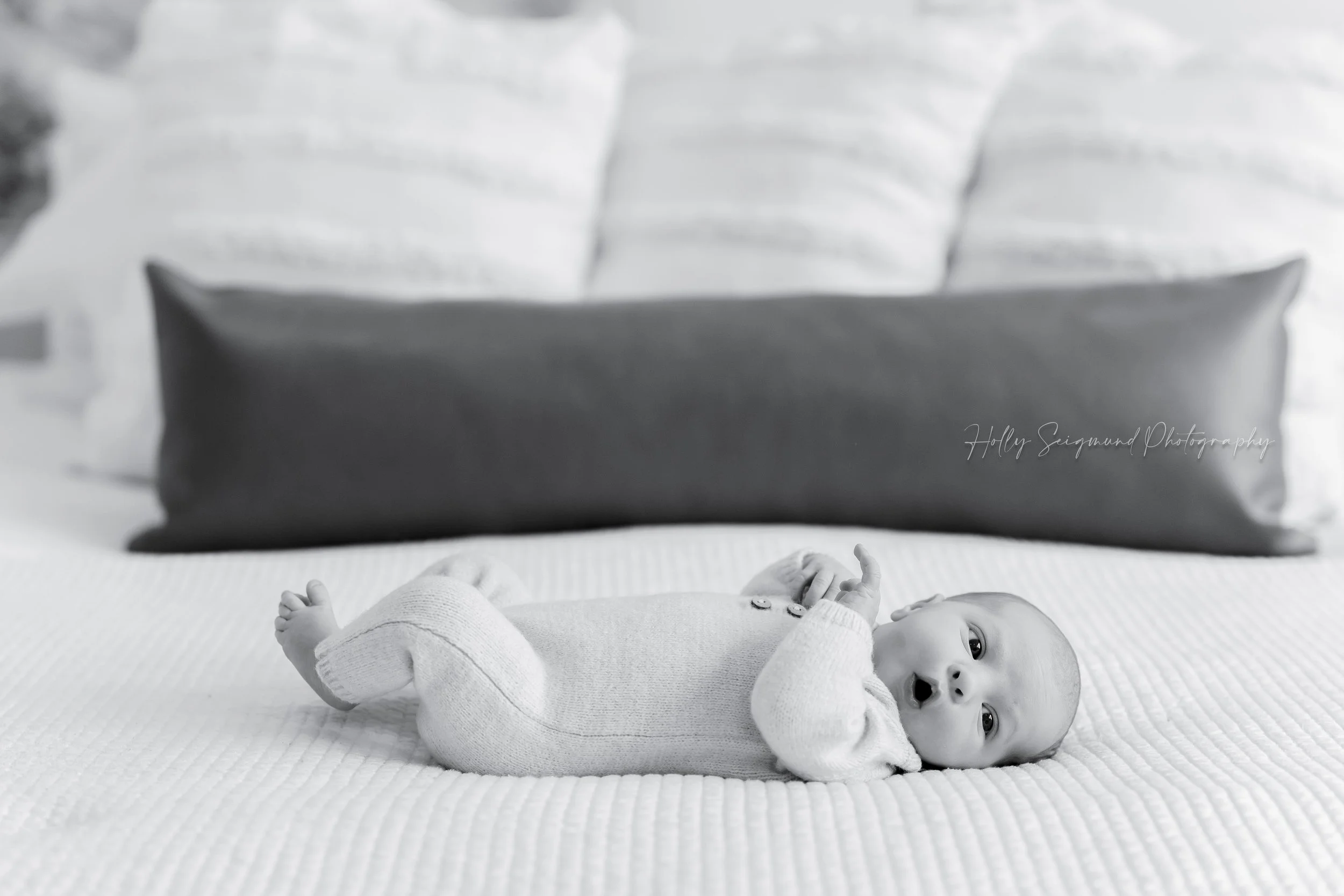 Black and white photo of a newborn baby dressed in a knit white outfit on a bed during a in-home newborn photography session in Dripping Springs, Texas