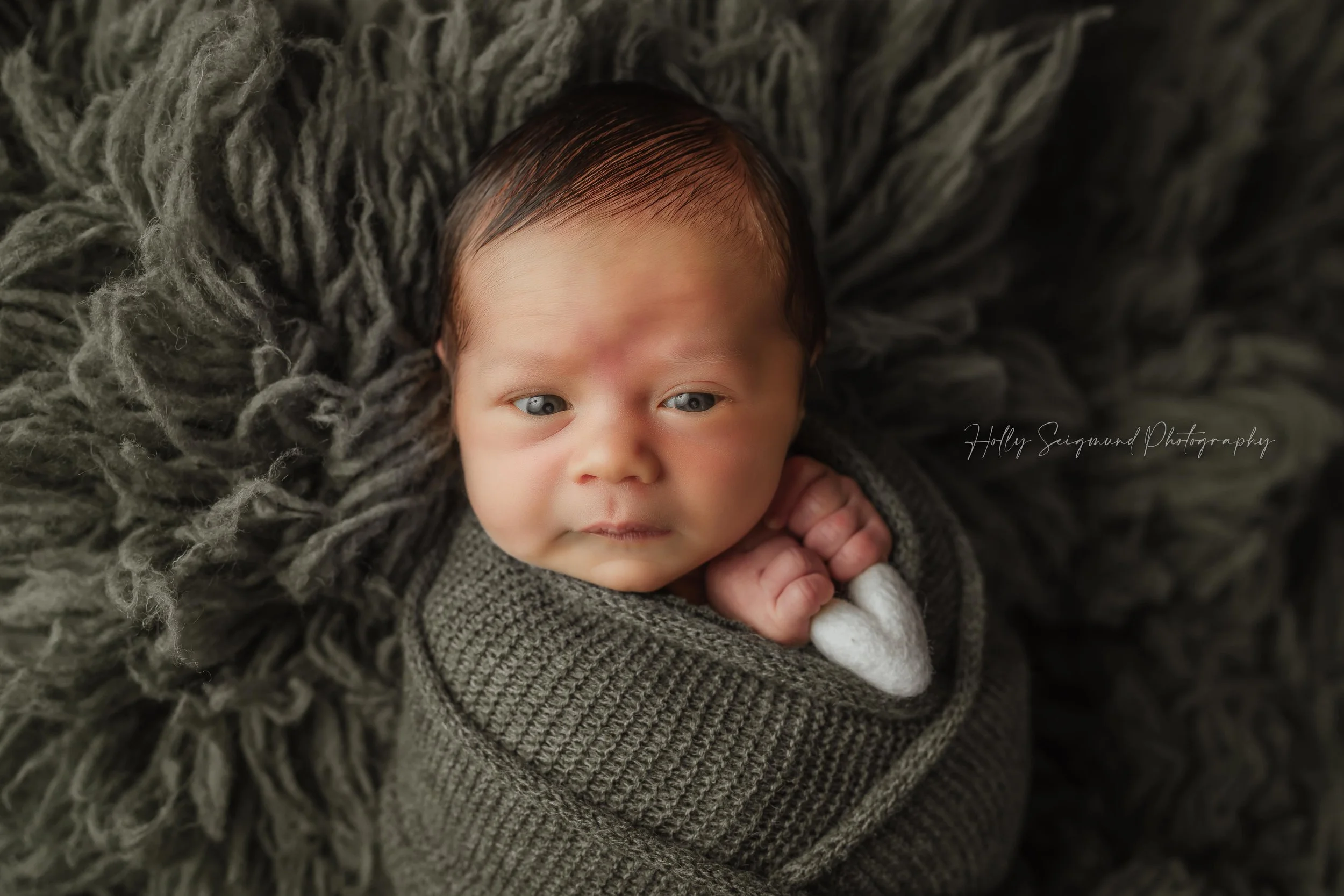 Baby boy with Olive green wrap and fluffy backdrop, holding a white felt heart during his newborn photography session in Blue, Texas