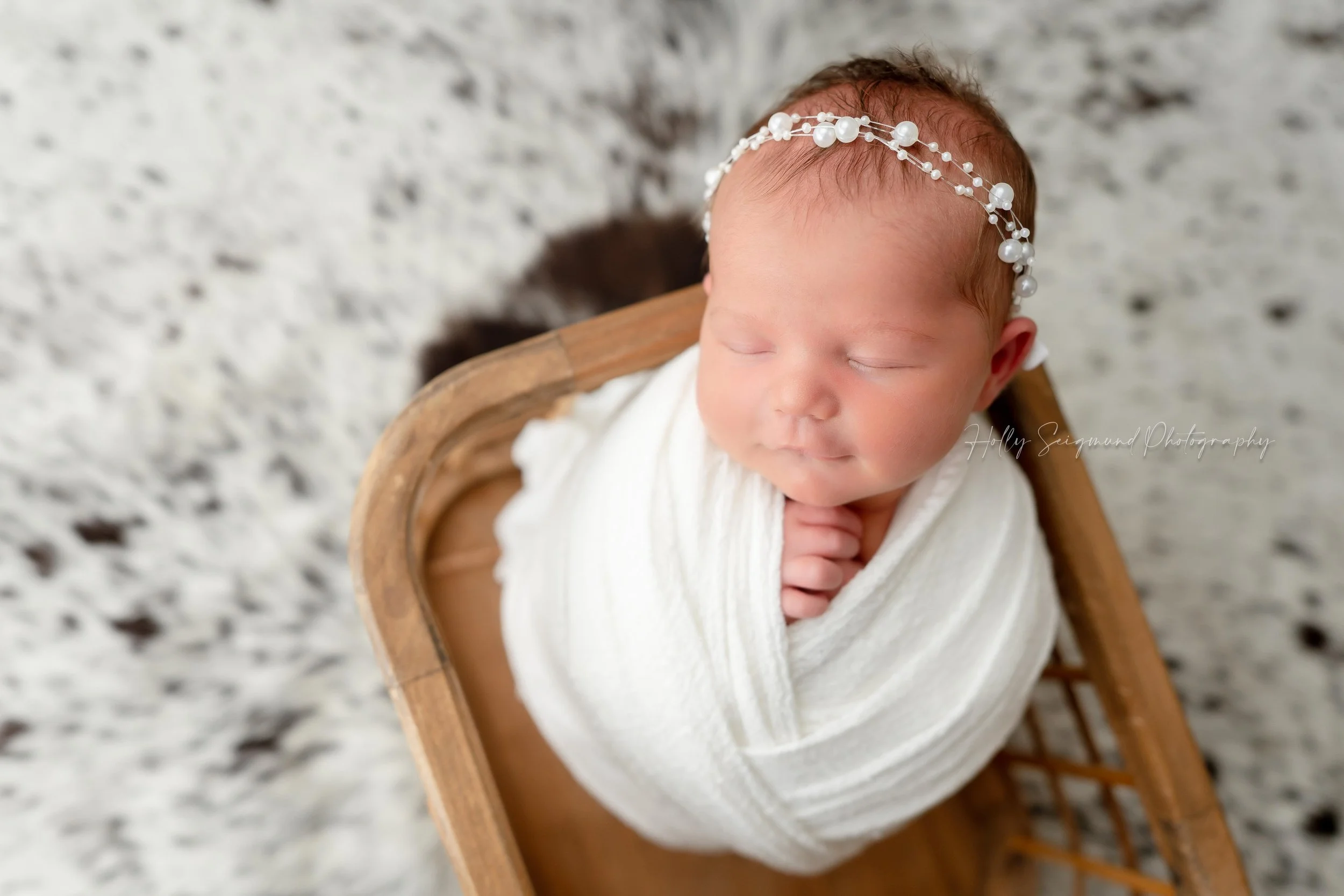 Western inspired newborn photography session with a baby girl wrapped in white with a pearl headband, sitting in a wooden crate and a cowhide backdrop in Lexington, Texas