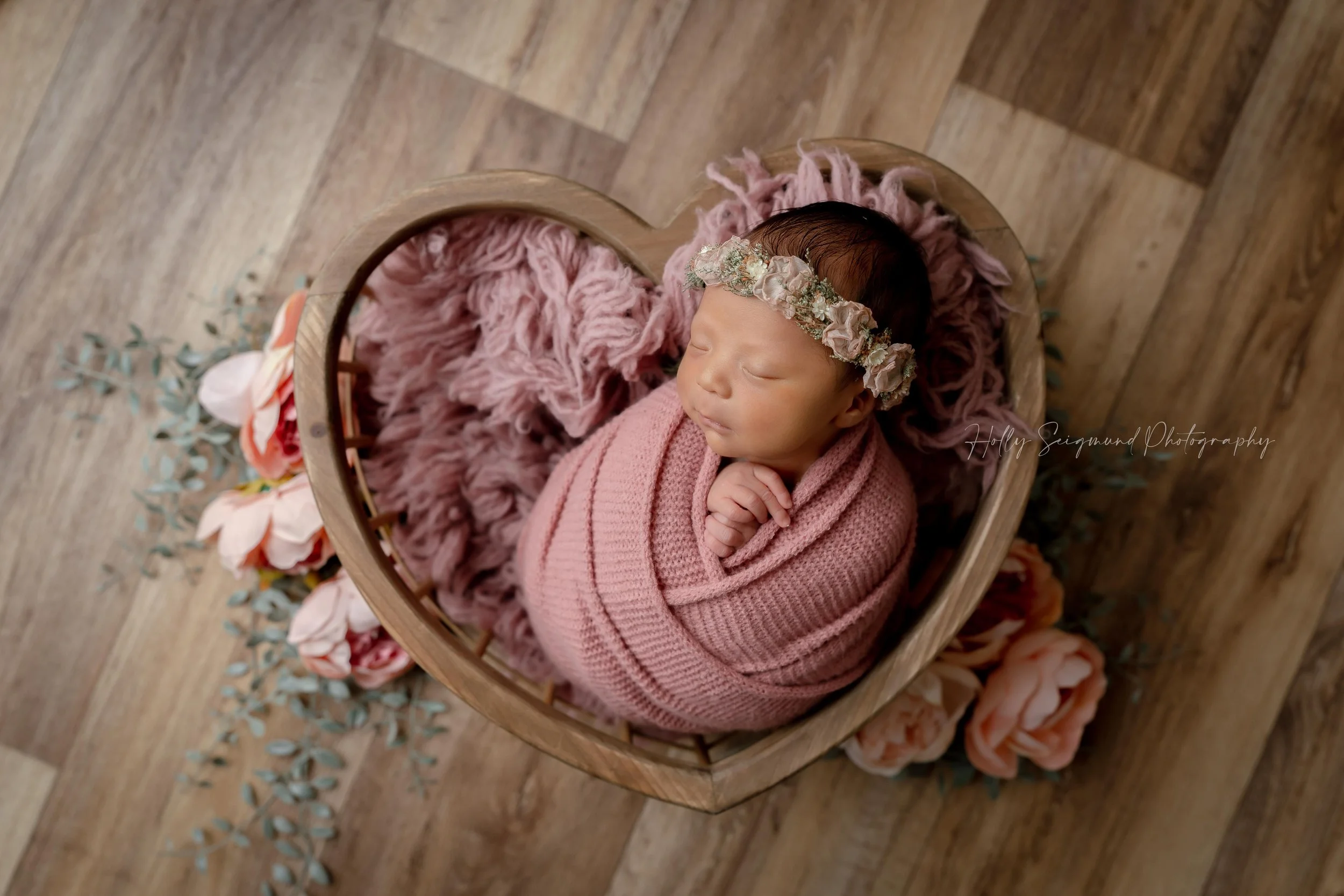 Wooden heart crate on a wood background with a newborn baby girl wrapped in pink with a floral crown in Lee County Texas